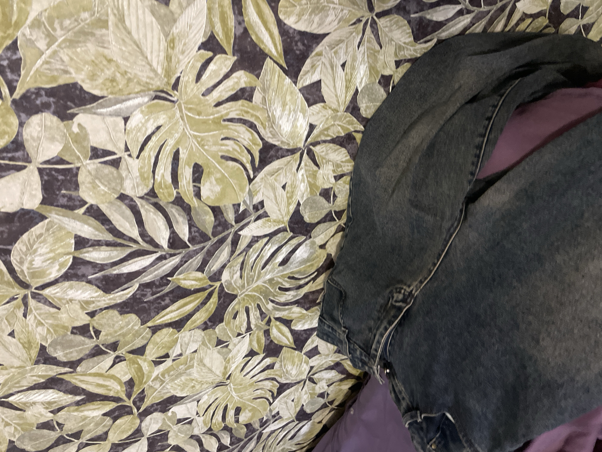 The image depicts a portion of a bed covered in patterned sheets with a leafy design. A folded denim jacket rests on the bed, partially covering a purple fabric item underneath. The location is not apparent from the image, and there is no indication it is in Olginka, Russia. No people or actions are visible.
