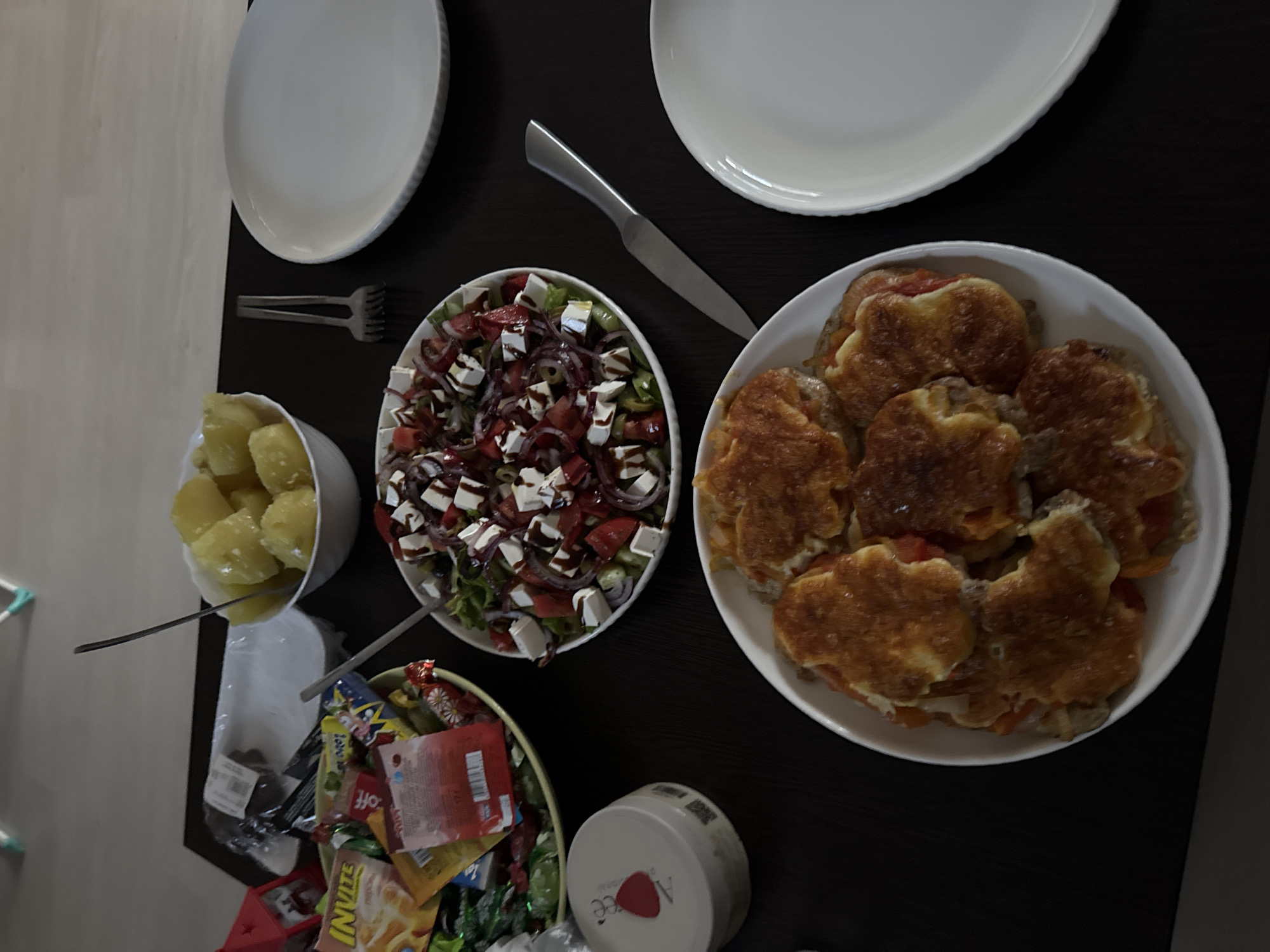 A top-down shot shows a table laden with food. In the foreground, a white bowl overflows with several pieces of golden-brown food, appearing to be patties of some kind, perhaps meat or vegetable based, topped with melted cheese and visible pieces of tomato. To the left and slightly above this bowl is another white bowl containing a fresh salad. The salad is a mix of greens, diced tomatoes, red onion rings, olives, and cubes of white cheese, possibly feta, drizzled with a dark dressing.
Above the salad bowl, a white bowl holds a generous serving of boiled potatoes, cut into large chunks. To the left of the salad and potatoes, a shallow, light-colored bowl is filled with an assortment of wrapped candies and snacks, including items labeled "INVITE". A small white container with a red heart logo and some text is visible near the candy bowl.
On the right side of the table, two plain white plates are stacked or placed next to each other. A silver fork rests between the potato bowl and the rightmost plate, and a silver knife lies between the salad bowl and the lower plate. The table surface is dark and appears to be made of wood. The background is out of focus, showing a neutral-colored wall and a sliver of what might be furniture. There are no people visible in the image. The scene depicts a meal spread, possibly a family gathering or a casual dinner. There is no indication of the city of Sochi, Russia, within the image content.