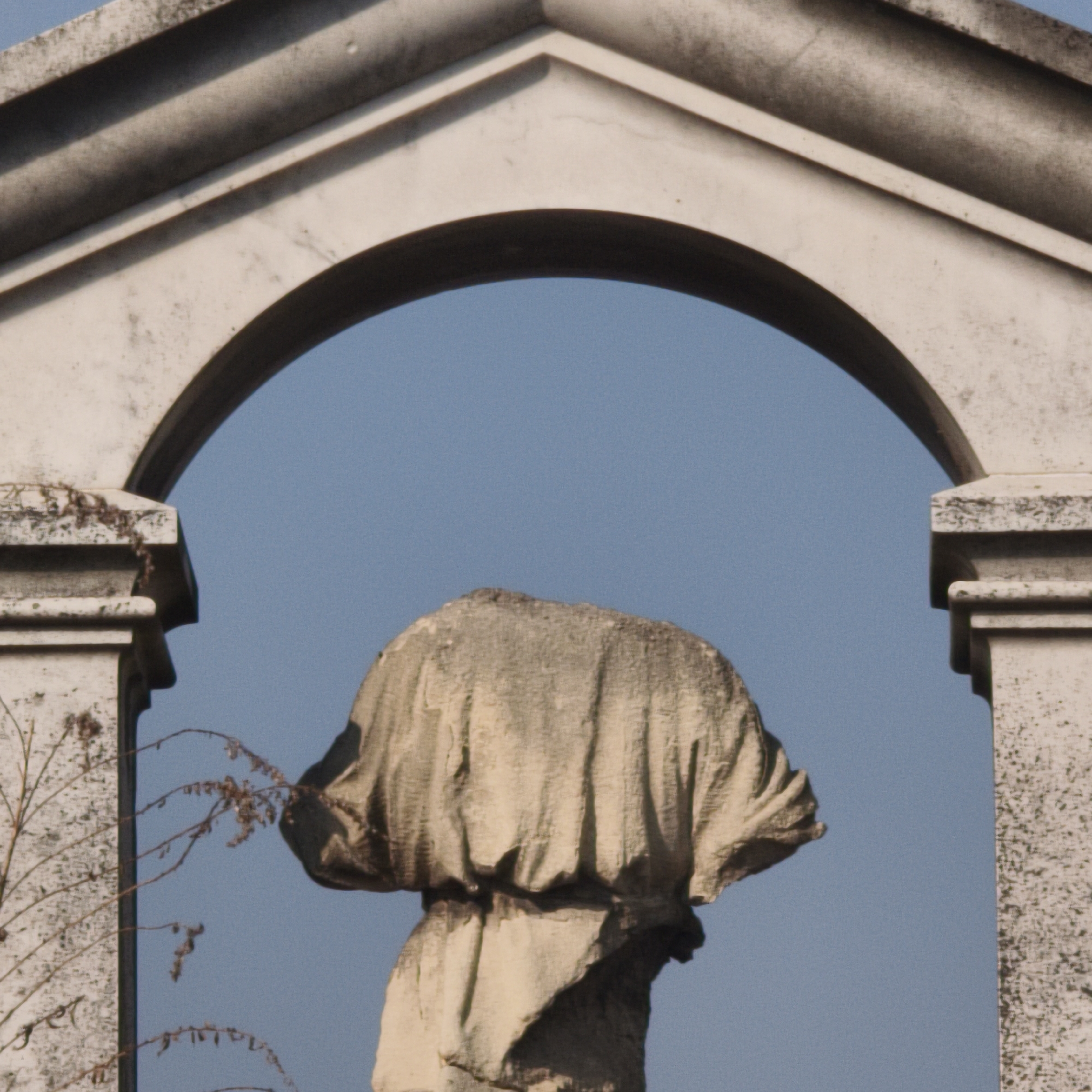 A weathered, headless stone statue is centrally positioned against a clear blue sky, framed by a light-colored, weathered stone archway. The statue depicts a draped figure, characterized by textured, deeply carved folds of fabric and a jagged, irregular top where the head is absent. The surrounding archway, featuring a rounded opening beneath a triangular pediment, exhibits significant surface discoloration, erosion, and minor biological growth, including dry, leafless plant tendrils clinging to the left vertical support. This scene is located in Hrodna, Belarus.