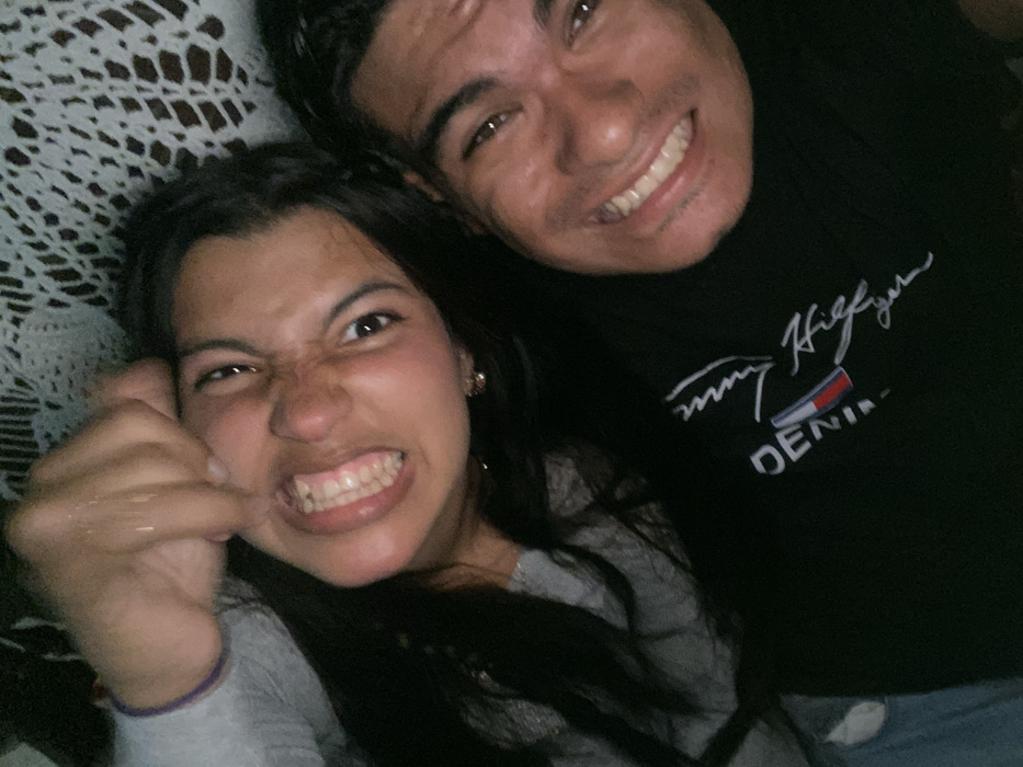 A young man and woman pose for a selfie. The woman, on the left, pulls her lip back to show her teeth while clenching her fist. The man, on the right, smiles widely. He wears a black Tommy Hilfiger t-shirt. The woman is in a gray long-sleeved shirt. The background includes what appears to be a white crocheted blanket. The overall lighting suggests the photo was taken indoors, possibly at night.