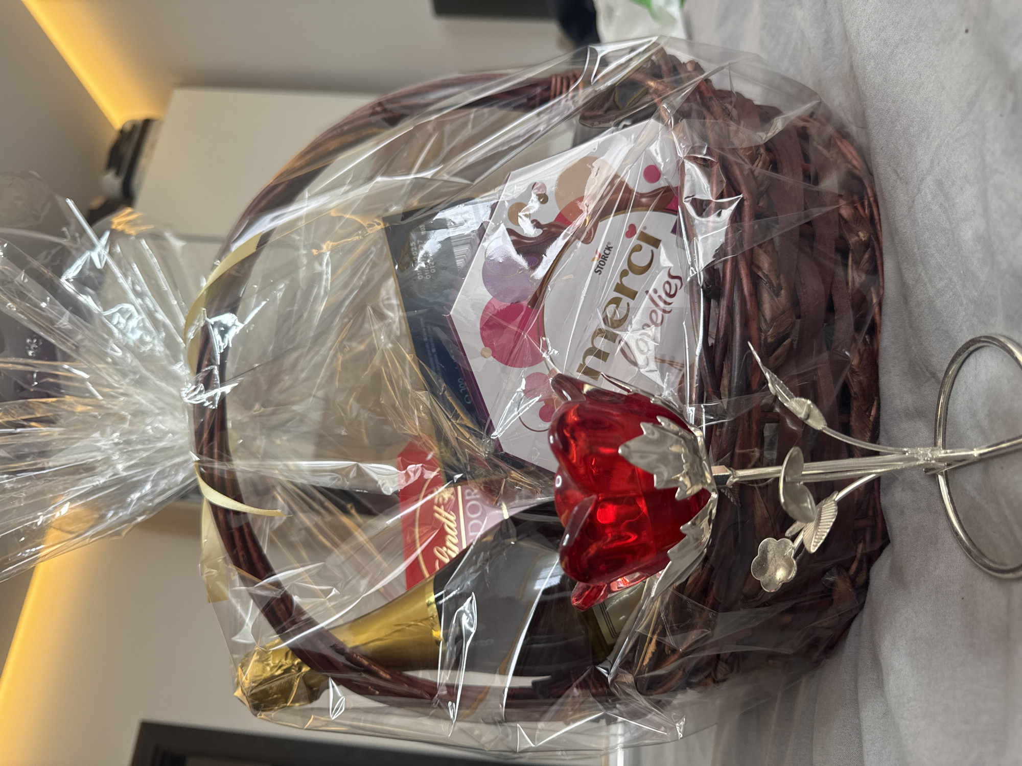 A gift basket is the central element of the image. It is wrapped in clear cellophane and contains various items: a bottle with a gold label, a box of Storck Merci chocolates, a Lindt chocolate item, and a metal rose with a red blossom. The basket itself is made of woven brown material. The basket is positioned on a light-colored surface with subtle textures suggesting a fabric tablecloth or bedsheet. In the background, out of focus, there is a glimpse of a room interior with light-colored walls and what appears to be a piece of furniture. There is no evidence to place the basket specifically in Sofia, Bulgaria.