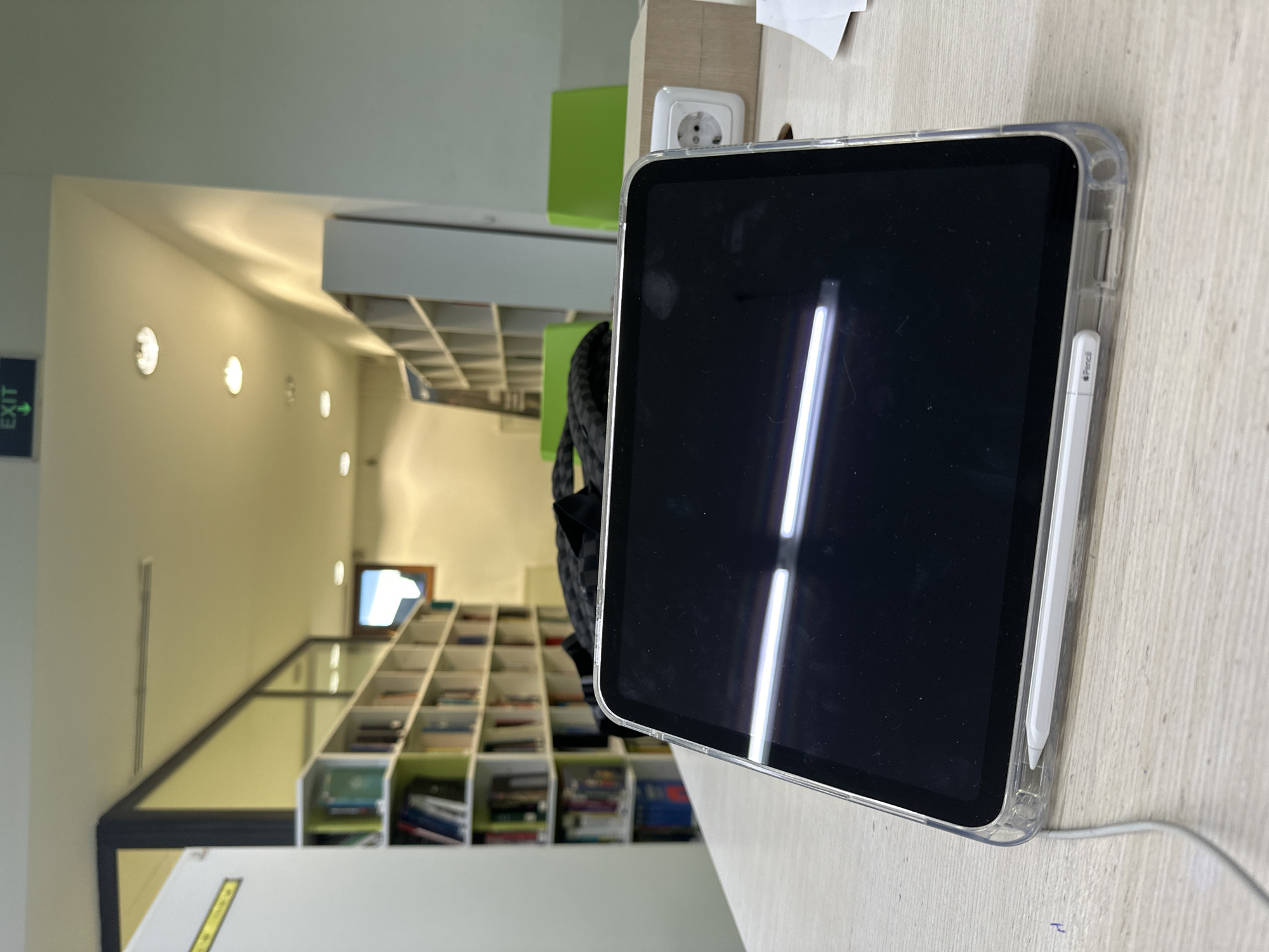 The image shows a library scene, likely in Yerevan, Armenia, given the context. A black and gray backpack rests near a light wood table. On the table, there is a covered iPad pro with a white pencil attached to the side. The library has rows of bookshelves filled with books and a few green chairs visible. There's an exit sign on the wall to the left, indicating the exit direction. There are multiple ceiling lights in the building. A white electrical outlet is present next to the iPad on the table. The image suggests someone is studying or working in the library, utilizing the iPad for their activities.