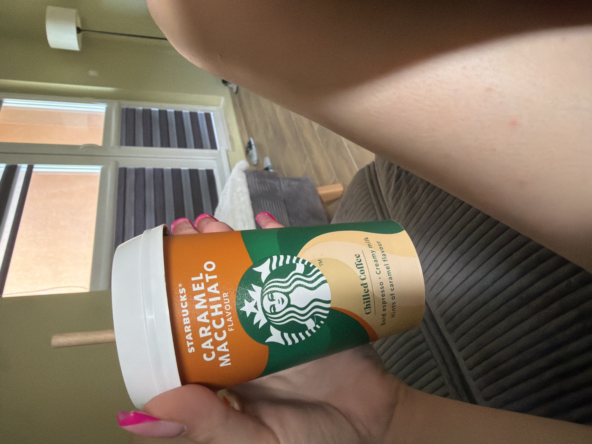 An adult woman is holding a Starbucks Caramel Macchiato chilled coffee in its orange and green branded cup, with a light brown straw protruding from the white lid. The woman's hands and nails, painted with bright pink nail polish, are visible on the left side of the frame, holding the cup. The background suggests an indoor domestic setting, likely a living room or bedroom, with a muted green wall, a large window with horizontal blinds, and a floor lamp with a white shade on the right. Part of a sofa or armchair, upholstered in dark grey corduroy, is visible in the foreground. A small red mark is present on the woman's exposed leg. No specific landmarks or elements definitively place the scene in Varna, Bulgaria.