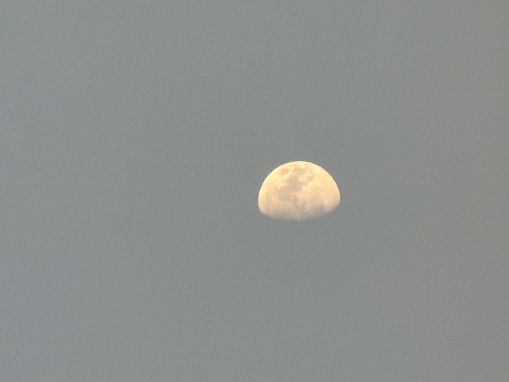 A photograph taken in the city of Kamyshin, Russia, shows a partially illuminated moon in a hazy, gray sky. The moon, appearing in a crescent phase, is visible slightly to the left of the center of the frame. Its surface exhibits some texture, indicating craters or shadows. The background is uniformly overcast with a muted gray tone, suggesting either dawn, dusk, or a heavily clouded daytime sky. There are no other celestial bodies, landforms, or man-made structures visible in the image. The lighting is soft and diffused, typical of an overcast environment.