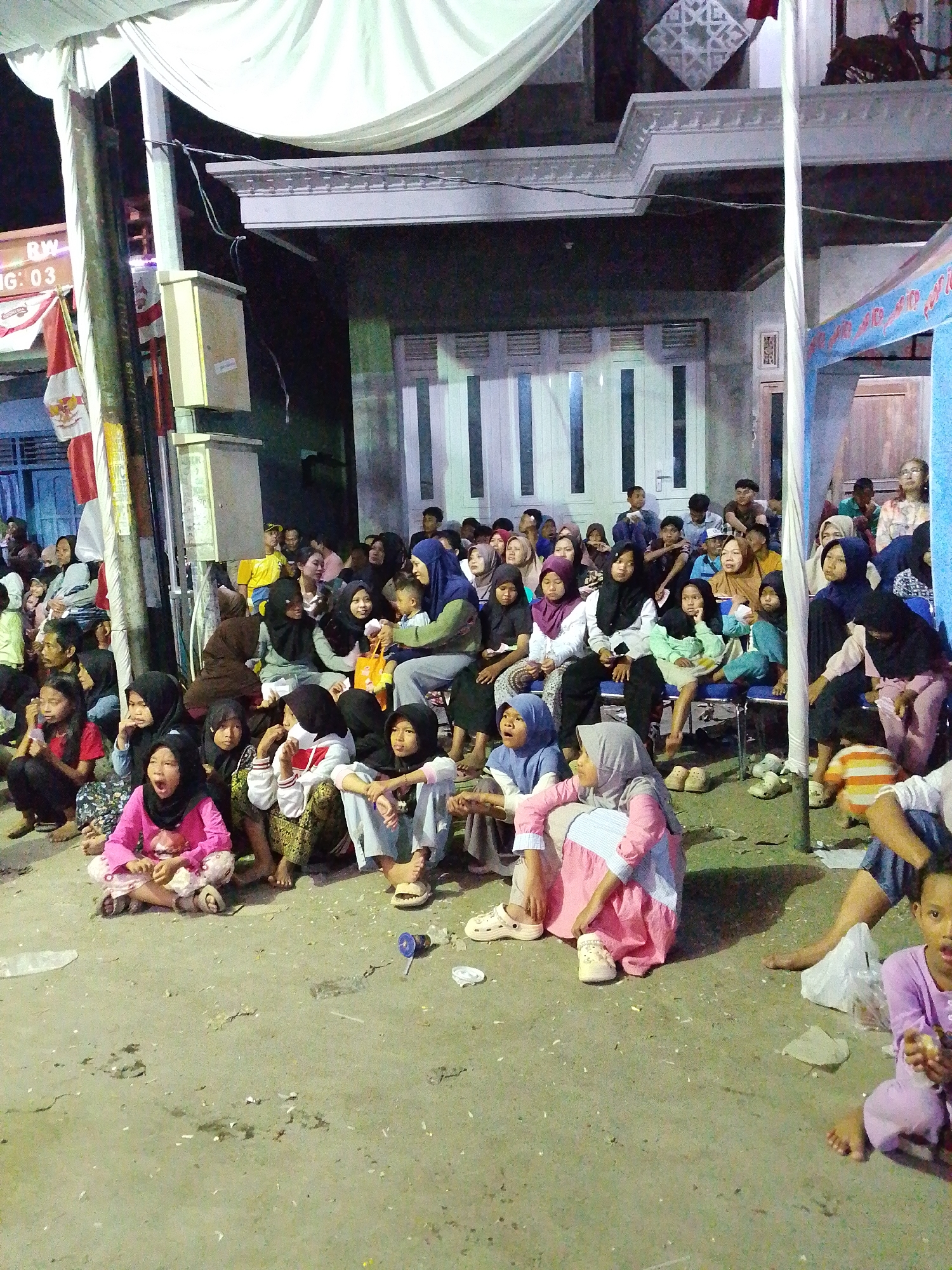 A group of people, predominantly women and children, are gathered outdoors, sitting on the ground and on chairs. They appear to be watching an event or performance, as many are looking in the same direction. The setting seems to be a temporary gathering space, possibly for a community event, with a white tent overhead and a building in the background. The ground is made of concrete or packed earth and is littered with debris. The overall atmosphere suggests a casual, communal gathering.