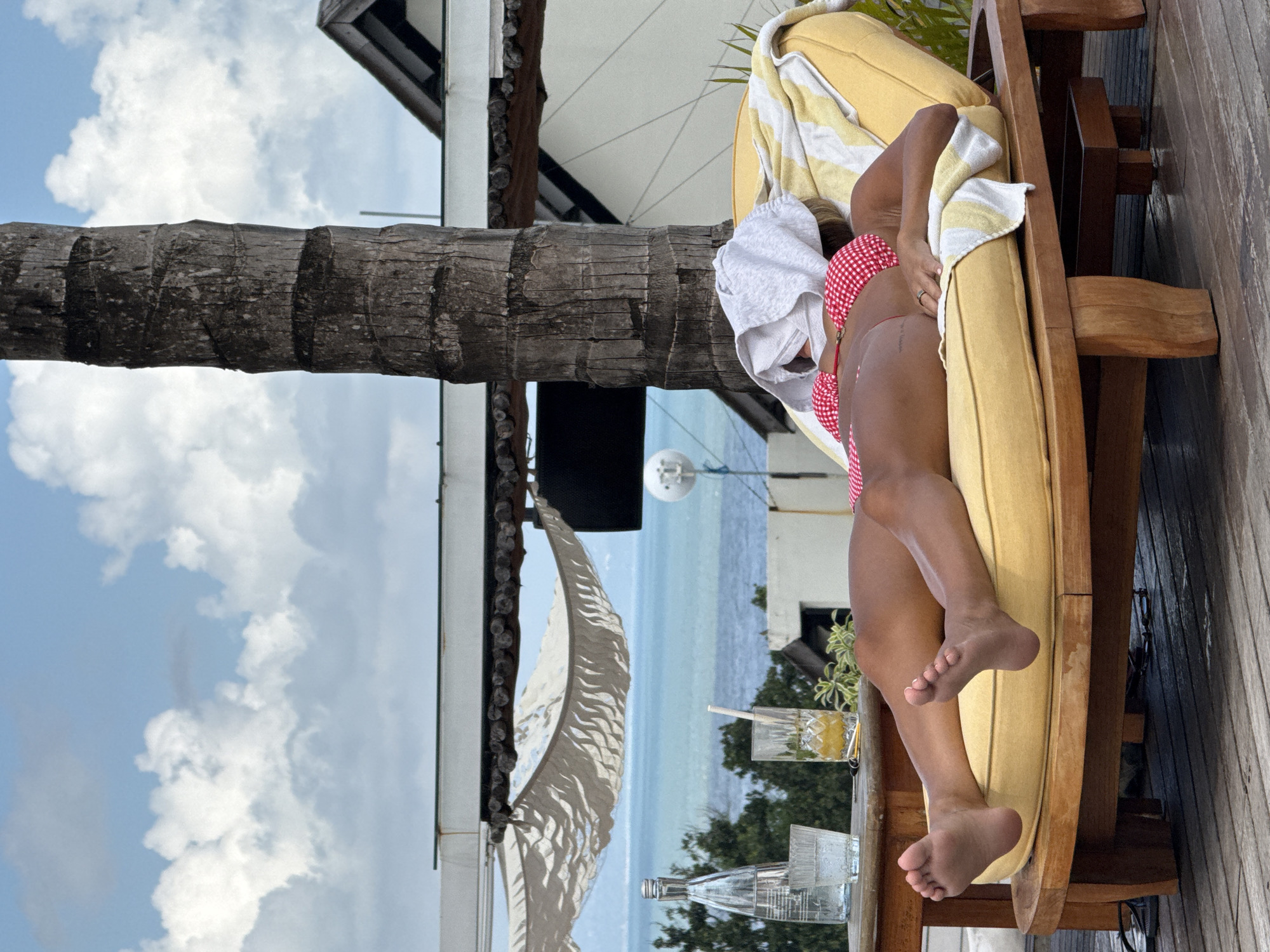 A woman is reclining on a yellow chaise lounge, with a towel draped over her head. She is wearing a red and white checkered bikini. There is a glass with a straw and a clear bottle of water on a table next to the chaise. The background features a palm tree, the ocean, and a white building. The scene appears to be outdoors in North Kuta, Indonesia.