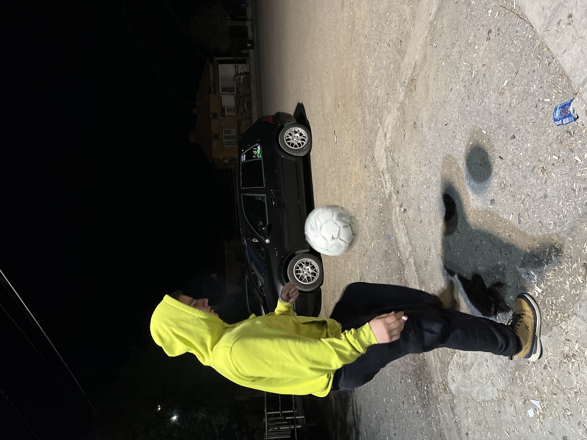A person wearing a neon yellow hooded sweatshirt, black pants, and tan shoes is juggling a soccer ball in front of a black car. The car is parked on a concrete surface that appears to be a road or parking area in Karavelovo, Bulgaria. The background shows buildings and dark sky, indicating it is nighttime. There's scattered debris on the concrete surface, along with a discarded beverage container.