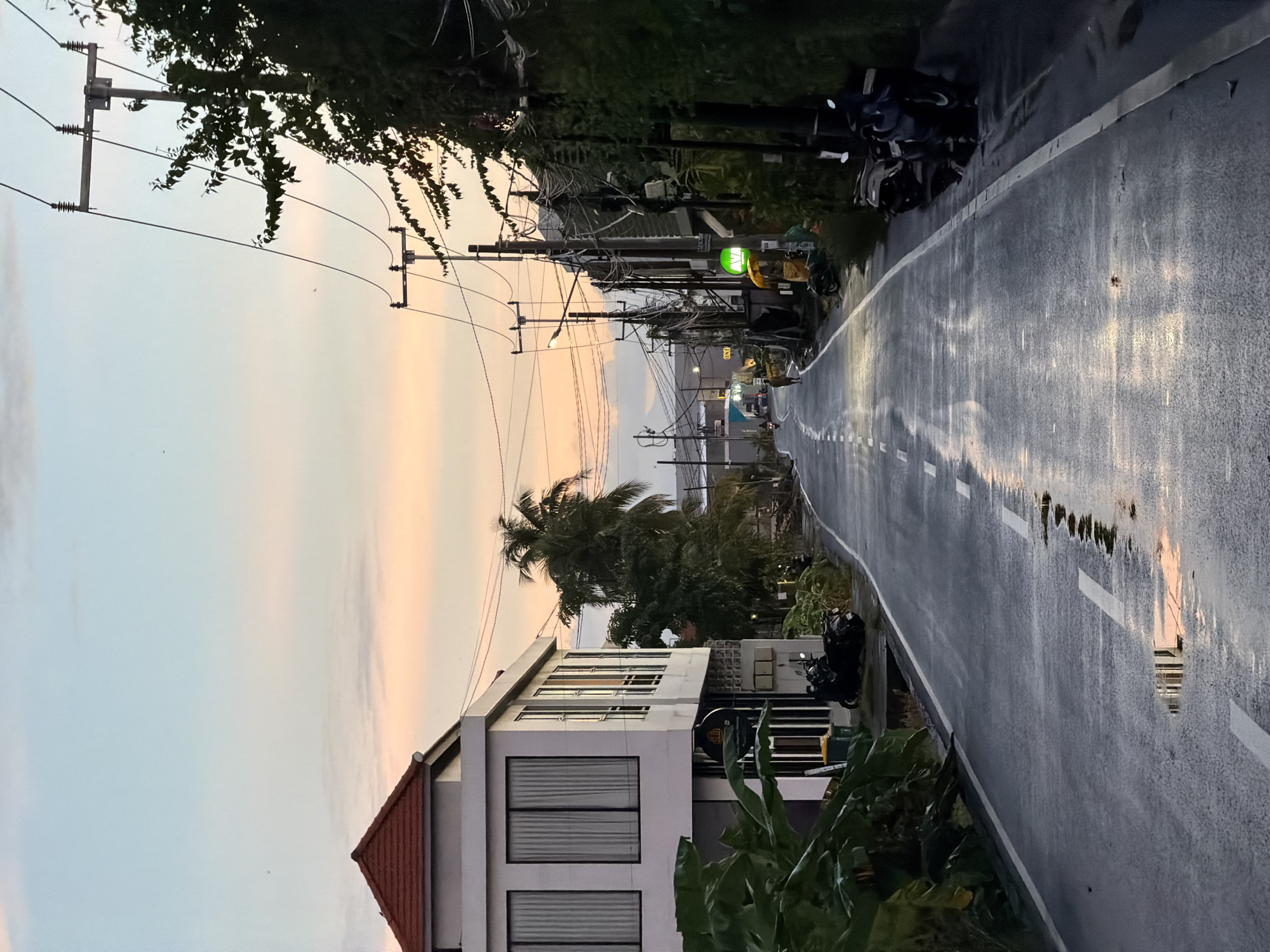 The image depicts a street in Mengwi, Indonesia, after a rain shower. The wet asphalt reflects the pastel hues of the sky at dusk. White lane markings are visible on the road. On both sides of the street are buildings and vegetation. Power lines run overhead, strung between utility poles. Several motorcycles are parked along the side of the road. In the distance, the ocean is visible.