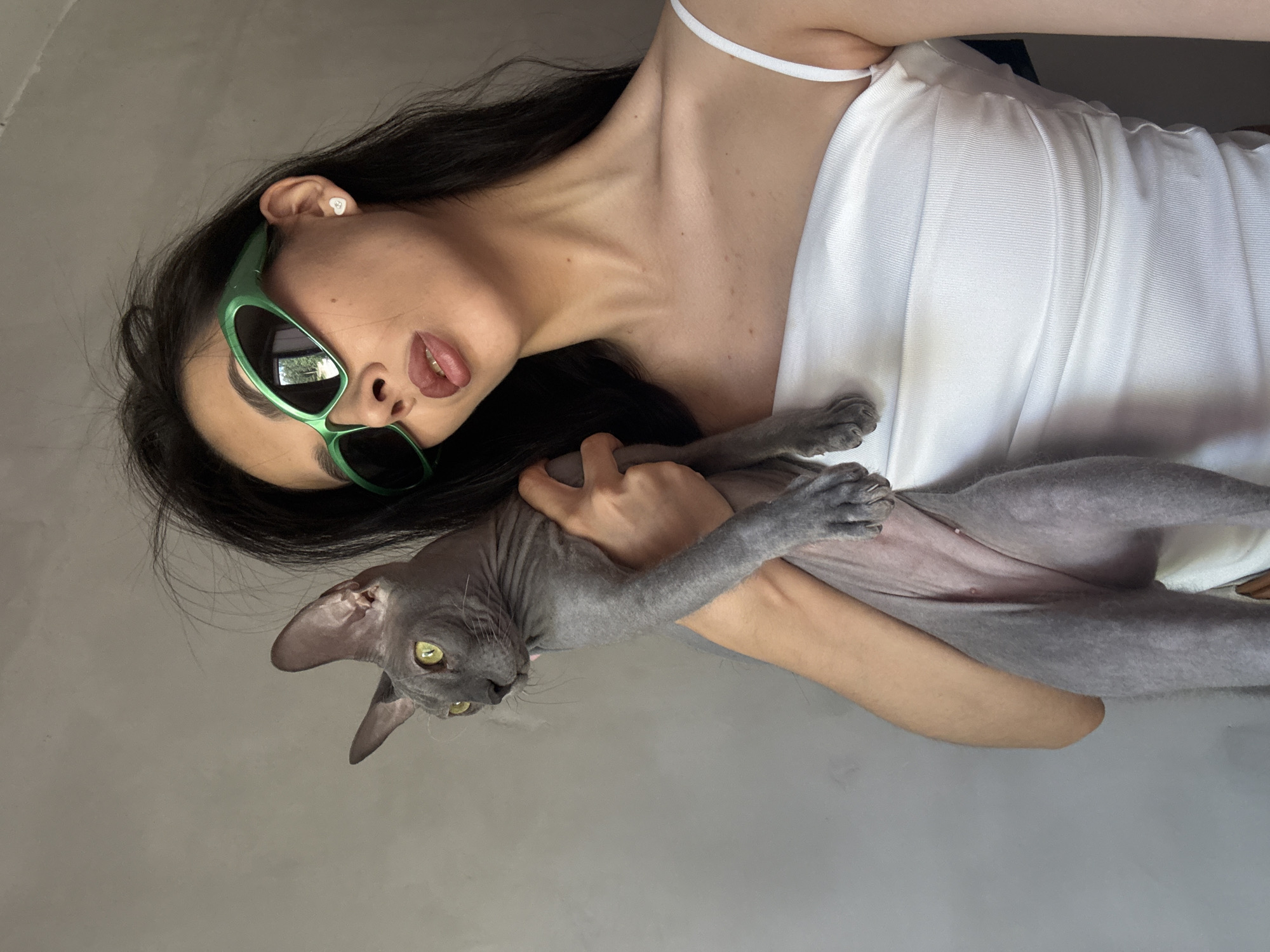A woman with dark hair and green sunglasses holds a gray Sphynx cat in front of a plain gray wall. She is wearing a white tube top. The cat is looking to the left. The woman is posing for a selfie, her face turned towards the camera. The background is a nondescript gray wall, providing no indication of a specific location within Votkinsk, Russia, or any other city.