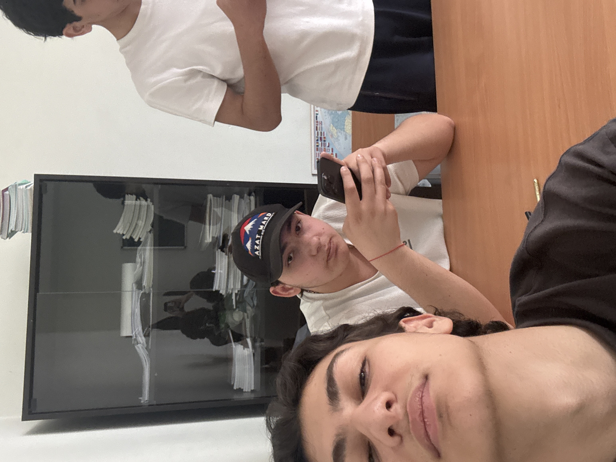 The image, rotated 90 degrees counter-clockwise, depicts three young males in an indoor setting. In the foreground, a male's face is partially visible from a low angle, showing his chin, mouth, nose, and part of his eyes, along with dark, wavy hair. He has a slight smile and is wearing a dark, long-sleeved top.
Behind him, a second male is seated, wearing a white t-shirt and a dark baseball cap with the visible text "AZAT MARD" on the front. He is holding a black smartphone with both hands, looking at its screen, while also glancing towards the camera. A red string bracelet is noticeable on his left wrist. His arm rests on a light brown wooden table or desk that occupies the right side of the frame.
Further in the background, a third male is partially visible from his chest up, wearing a white t-shirt and dark trousers. His head is turned away from the camera, facing the left.
To the left side of the image, a dark, glass-fronted cabinet or bookcase is filled with stacks of papers or books, showing reflections on its surface. A white wall forms the background for the male at the top of the frame, and a poster or map featuring numerous small rectangular patterns is visible on this wall behind the seated male's elbow. The lighting suggests it is daytime. The "AZAT MARD" text on the cap is a notable detail.