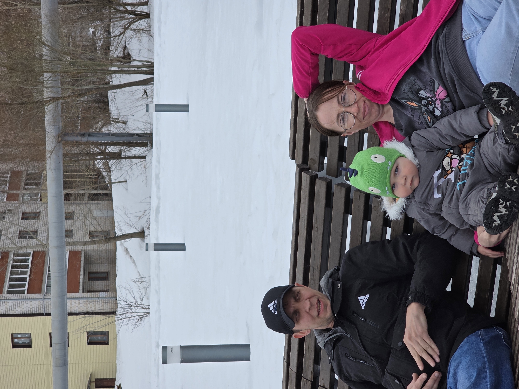 A family is sitting on a wooden bench in what appears to be the city of Votkinsk, Russia. Snow covers the ground around the bench and is visible in the background. A man, wearing a black Adidas jacket and cap, sits on the bench with his hand on his stomach. Beside him sits a young child wearing a green dinosaur hat and a grey jacket, also on the bench. A woman, wearing glasses, a pink jacket, and jeans, is positioned behind the child on the bench. The background includes buildings and bare trees, typical of an urban winter landscape. The image is rotated counterclockwise.