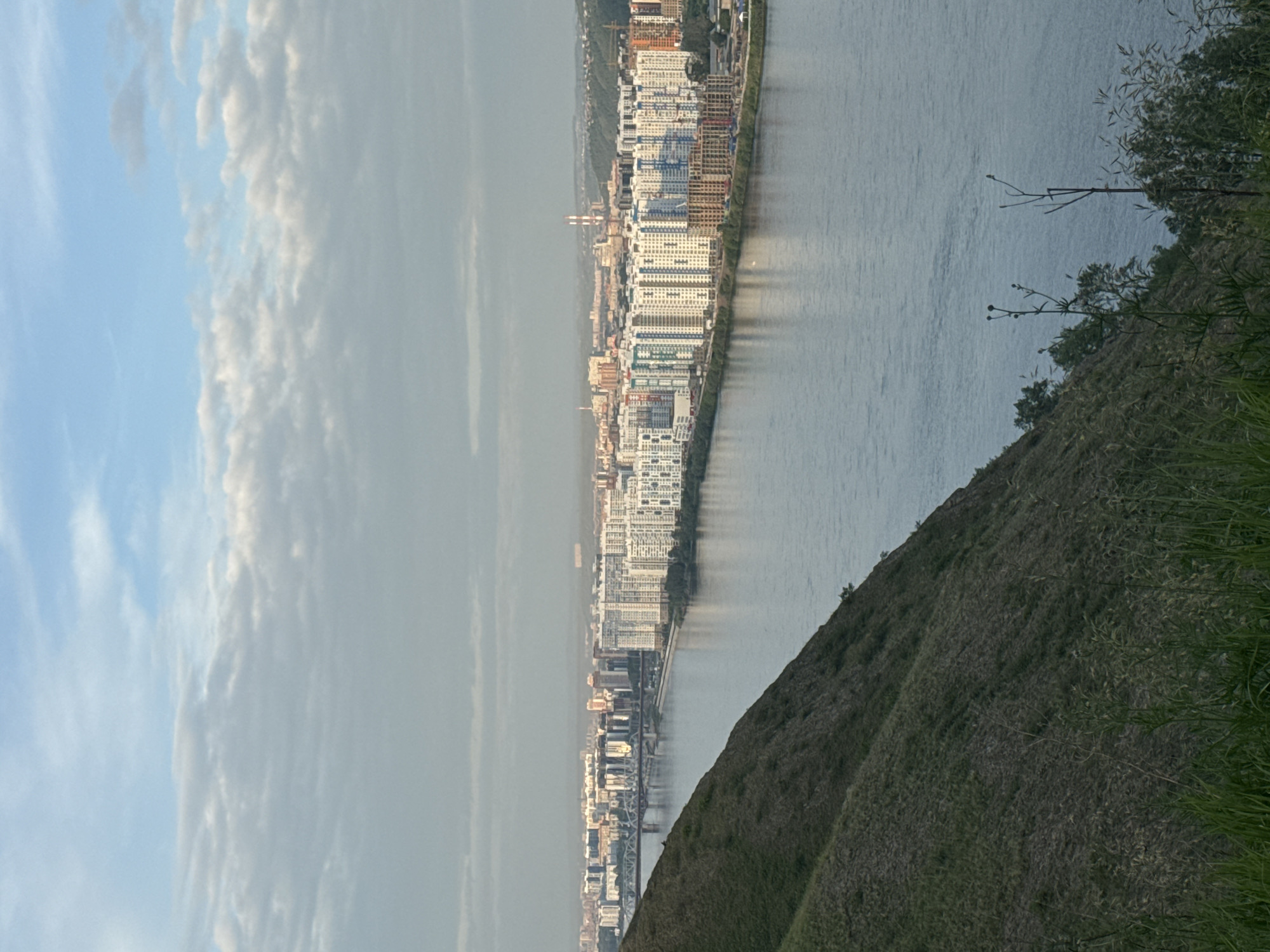 This is an outdoor, high-angle, landscape photograph of the city of Krasnoyarsk, Russia, featuring the Yenisei River and the city skyline. The image is dominated by the river in the foreground, reflecting the sky and the city lights. On the left side, a steep, grassy hill occupies a significant portion of the frame, sparsely covered with vegetation. Across the river, the city of Krasnoyarsk stretches along the opposite bank, characterized by a dense collection of modern, multi-story residential buildings with white and colorful facades. A bridge spans the Yenisei River, connecting the two sides of the city. The sky above is overcast with a mix of grey and white clouds, with hints of blue peeking through. The overall lighting suggests it is either early morning or late evening, with a soft, diffused light illuminating the scene. There are no people or animals visible in the image.