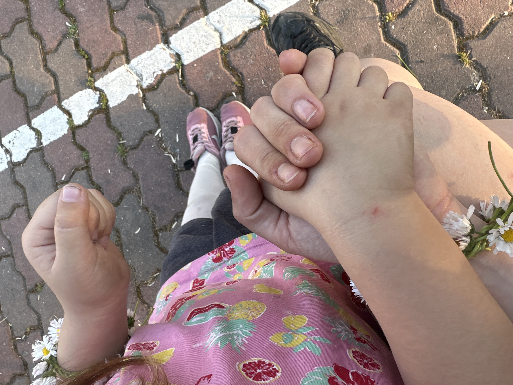 A high-angle shot of two people holding hands. One person has pale skin and is wearing black sneakers. The other person, presumably a child, is wearing pink sneakers, white leggings, and a floral patterned pink dress. The child's hand is clenched in a fist on the left. Both people are on a brick or concrete sidewalk with white stripes. A daisy chain is on the child's arm. The image appears to have been taken outside in daylight. The location is not specified, other than Resen, Bulgaria.