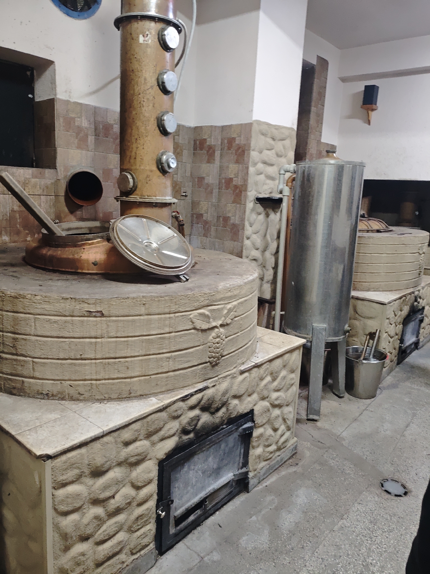 The image captures the interior of a distillery. The scene is dominated by a large, cylindrical, copper still, which is the primary piece of equipment for distilling spirits. This still is seated atop a brick-like stone base that appears to have a decorative grape pattern carved into its side. A metallic lid is partially open, revealing the interior of the still. To the right of the still is a tall, stainless steel tank, which also appears to be a part of the distilling apparatus. Beside the tank, a metal bucket holds various tools or utensils. The walls are partially covered with textured, stone-like panels and tiled sections. A dark opening is visible behind the stainless steel tank, likely a fireplace. The floor is made of concrete. The setting appears to be an artisanal or small-scale distillery. The specific city and location are not discernible from the image. There are no people visible in the scene.