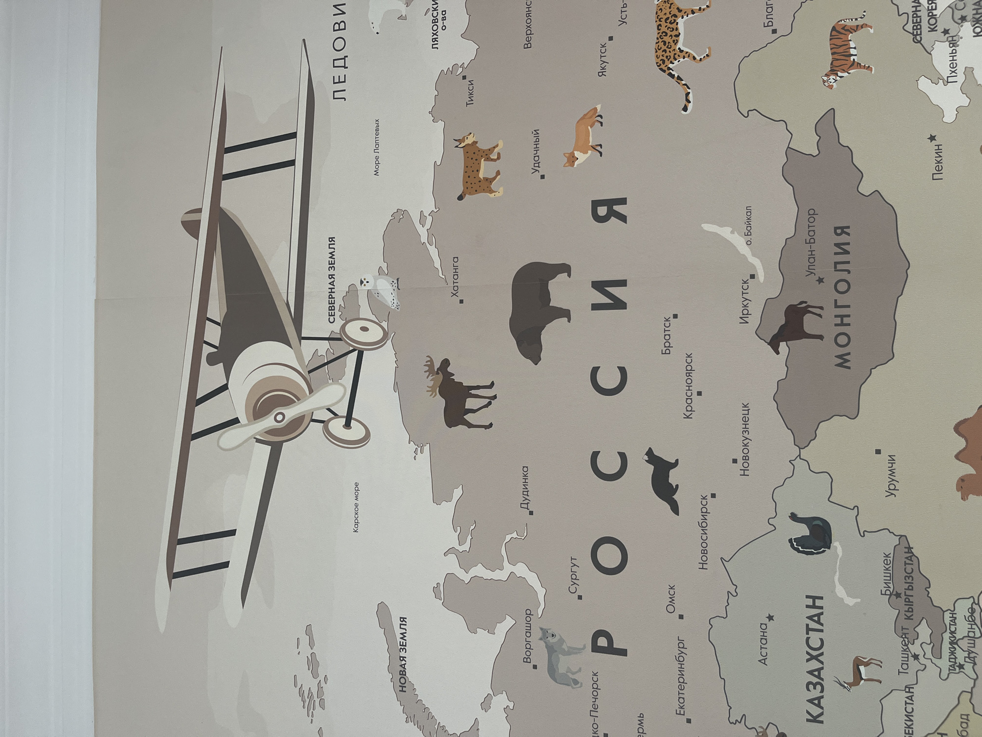 The image is a close-up of a world map, likely a children's educational map. The map features illustrations of animals corresponding to their native regions. A vintage biplane illustration is overlaid on the map. Place names are written in Cyrillic script. The prominent land masses depicted are Russia, Mongolia, and Kazakhstan. There is no indication of Kaspiysk or its location.