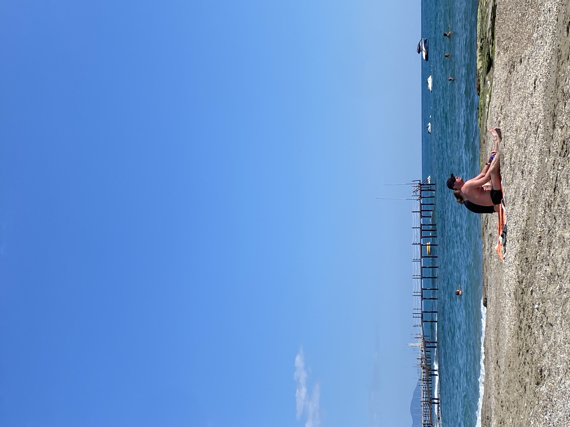 The scene is a beach in Alanya, Turkey, where a man sits on the sand with his legs crossed, wearing black swim trunks and a cap. An orange and black towel lies beneath him. Behind him is a shoreline littered with small shells. The ocean stretches out to a wooden pier with several vertical supports and horizontal railings. Further out, several people are swimming in the ocean, and a few boats are visible on the horizon. The sky is clear and blue with a few small clouds near the bottom left. The overall impression is a sunny day at the beach.