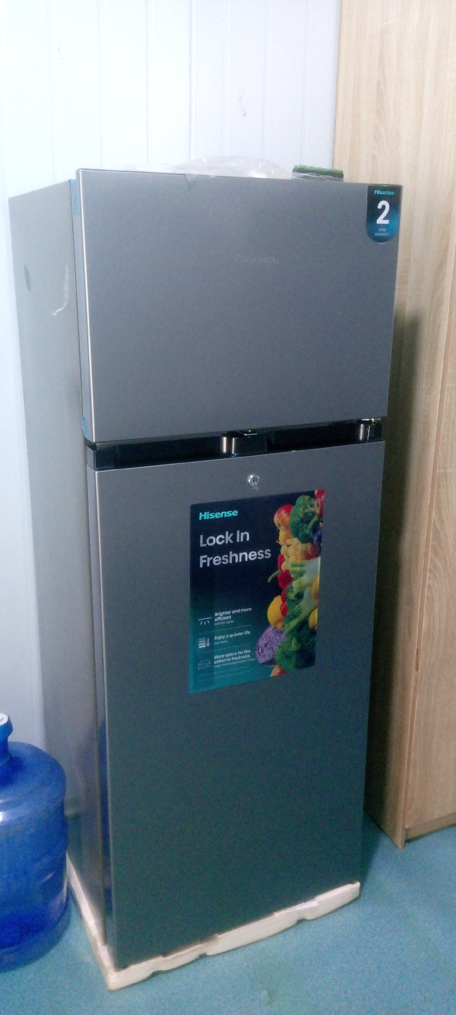 A silver Hisense refrigerator with a "2 Year Warranty" sticker is positioned in a room. The refrigerator has a top freezer compartment and a bottom refrigerator compartment. A sticker on the refrigerator door reads "Hisense Lock In Freshness" and displays images of fruits and vegetables. To the left of the refrigerator, a blue water jug is partially visible. The room has light-colored walls and a blue floor. The scene is likely indoors in a residential or commercial setting in Indo, Nigeria.