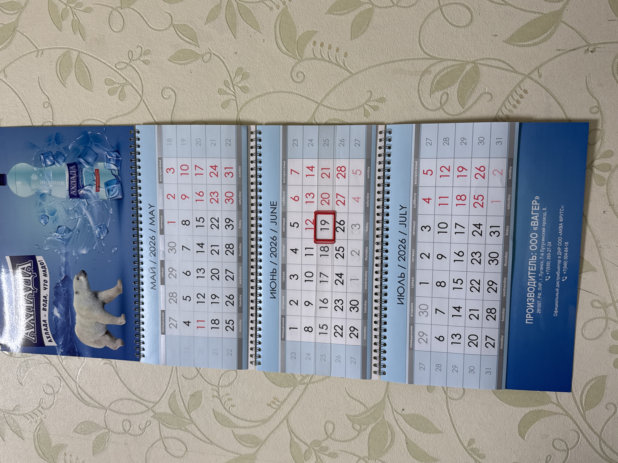 This media file is a close-up shot of a wall calendar featuring an image of a polar bear beside a bottle of "Akhlada" mineral water. The calendar pages visible are for May, June, and July of 2026. The May page features the polar bear image and advertising text for "Akhlada" water. The June and July pages are typical calendar grids with days of the week and dates. The calendar also includes contact information for a manufacturer and distributor. The overall scene suggests a commercial or promotional context, possibly for the "Akhlada" brand. The lighting and wallpaper background indicate the photo was taken indoors, likely in a home or office environment.