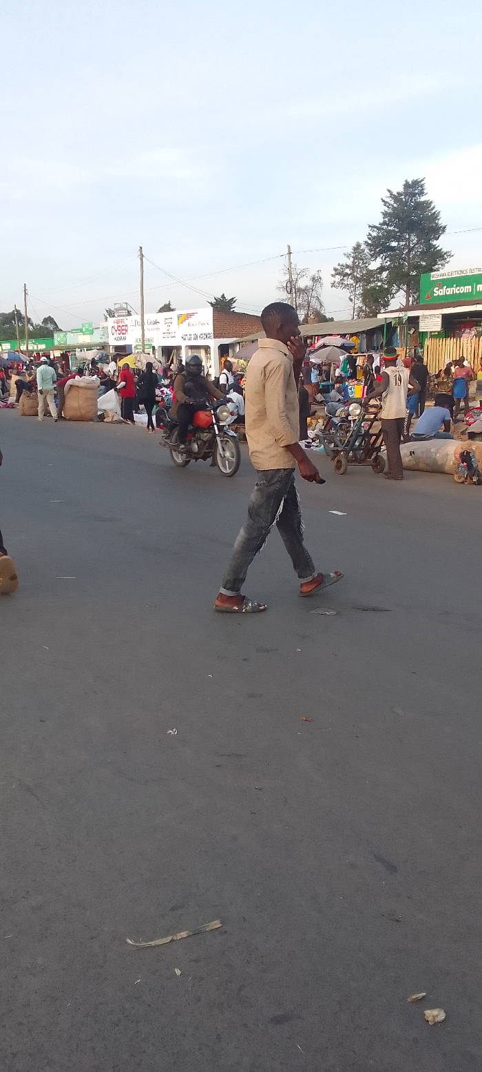 This image depicts a bustling street scene in Kakunga, Kenya, likely during the late afternoon or early evening hours under a bright, slightly hazy sky. The central figure is a young man walking across the paved road from right to left, holding a mobile phone to his ear. He is dressed in a light-colored, long-sleeved collared shirt, faded ripped blue jeans, and open-toed sandals worn over orange socks.
The background reveals a lively market area with numerous people, market stalls, and shops lining both sides of the street. On the left, several individuals are gathered around large sacks and piles of goods. A person in a light blue shirt is visible among them. In the mid-ground, a person wearing a dark helmet and jacket rides a red and black motorcycle, heading left. Further along the street on the right, more people are seen near market stands, some covered by umbrellas.
Commercial buildings with various signs flank the road. Visible text includes a prominent green "Safaricom m-pesa" sign, a "CYBER" sign, and "MESHANA ELECTRONICS ELECTRICAL" on another sign. Overhead power lines and utility poles are present. Trees are scattered behind the buildings. The road itself is asphalt, with some minor debris visible on its surface. The scene captures a dynamic moment of daily life and commerce in a busy town.