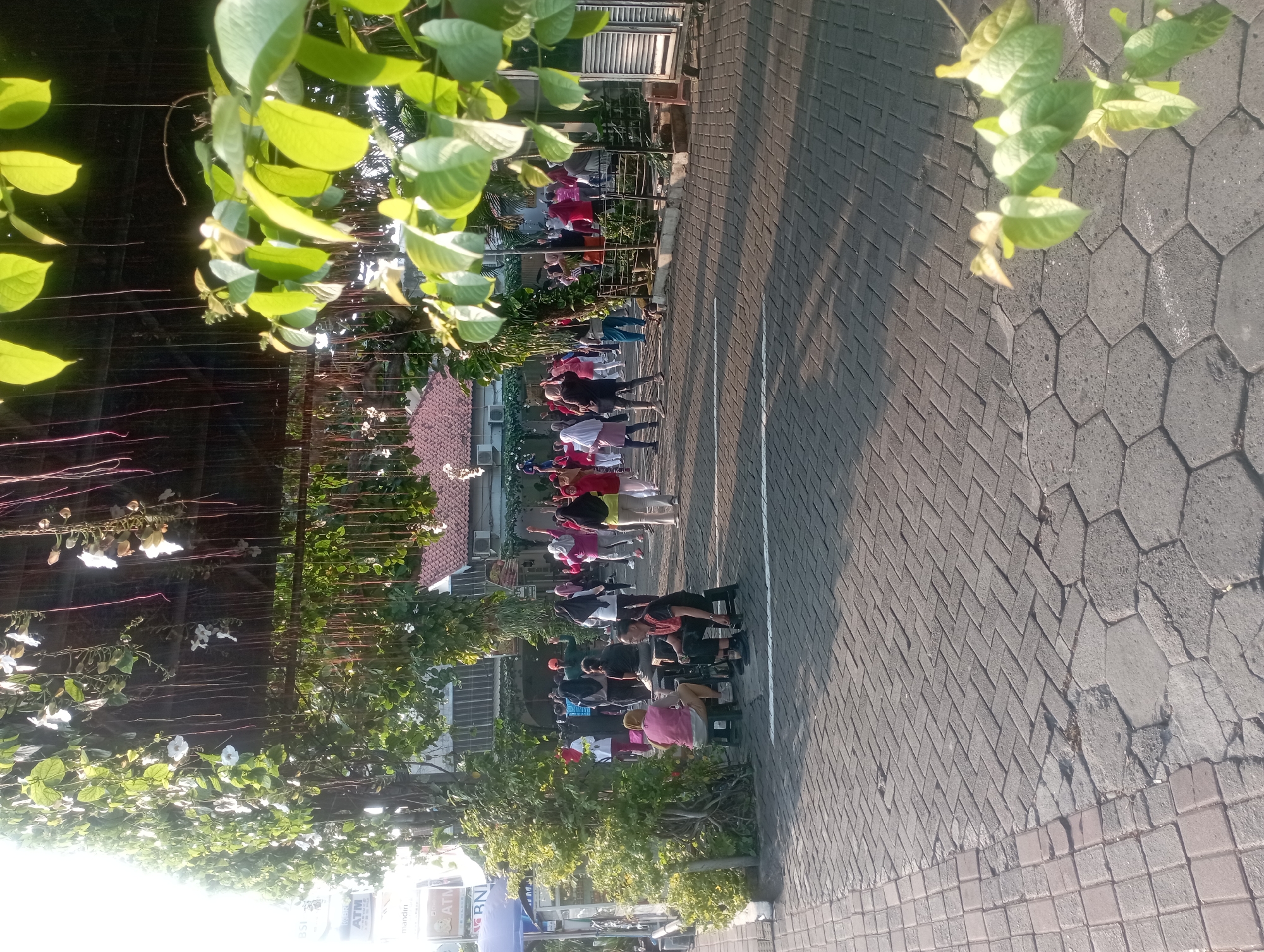 An outdoor scene shows a group of people exercising in a paved area in front of a building. The building appears to be a commercial establishment, with signage visible, including "BNI" and "ATM." Lush green vines with white flowers cascade from an overhead structure, casting shadows on the ground. The ground is paved with interlocking bricks. Some individuals are actively participating in exercises, while others are seated on small stools, observing or resting. The lighting suggests it is either early morning or late afternoon. The overall atmosphere is one of community activity in a semi-urban setting. The scene is identifiable as being in the city of Yogyakarta, Indonesia, due to the specific ATM branding and architectural style.