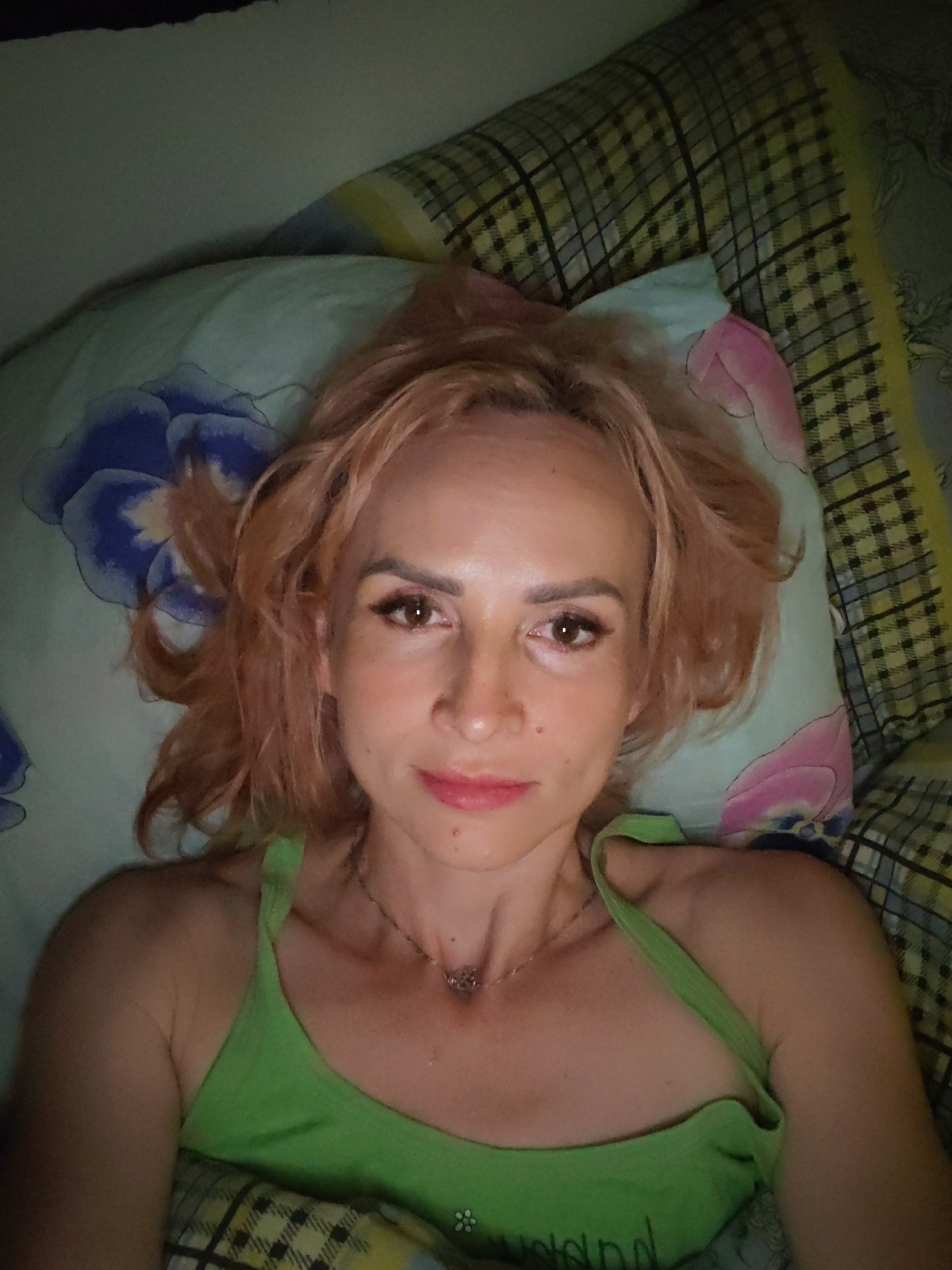 A woman with light brown, shoulder-length hair is lying on a pillow in bed. She is looking directly at the camera. She is wearing a green tank top. The pillow has a floral pattern with blue and pink flowers. The background shows a plaid blanket. The lighting appears to be artificial. The image was taken in Shalsky, Russia.