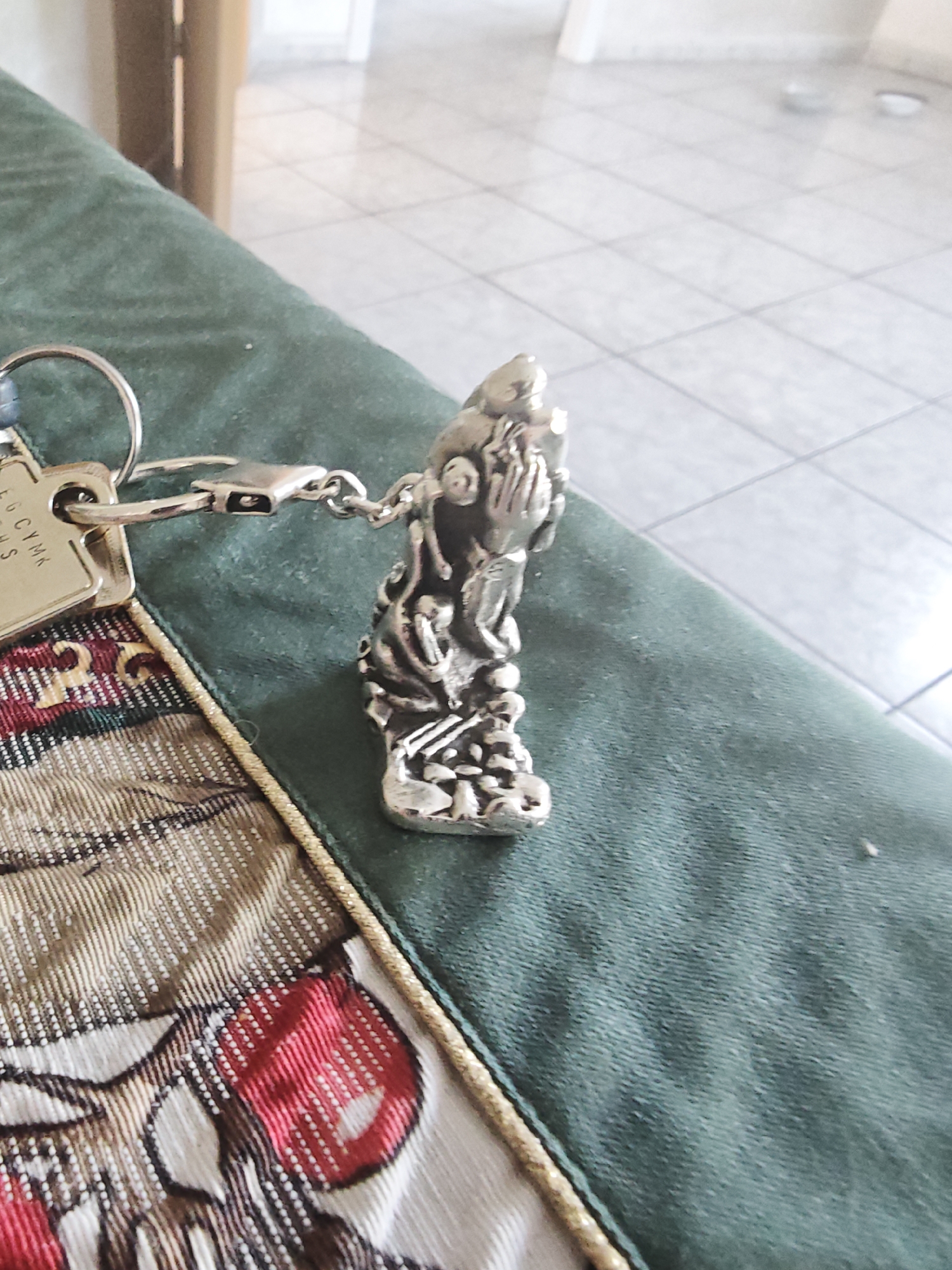 A silver keychain adorns a dark green textured surface, likely upholstery or fabric. The keychain features a small, intricate charm with a figure that appears to be praying or supplicating, with hands clasped. The charm is connected by a delicate chain to a clasp, which in turn is attached to a circular keyring. Also attached to the keyring is a small, rectangular tag. Faint text is visible on the tag, appearing to be a series of letters or a code, partially obscured and difficult to read precisely. The overall impression is that of a personal item, perhaps a lucky charm or a memento, resting on a surface that suggests an indoor setting. The background is blurred, showing tiled flooring and hints of vertical structures, possibly furniture or doorways, implying a domestic or commercial interior. No specific time of day or weather conditions are discernible.