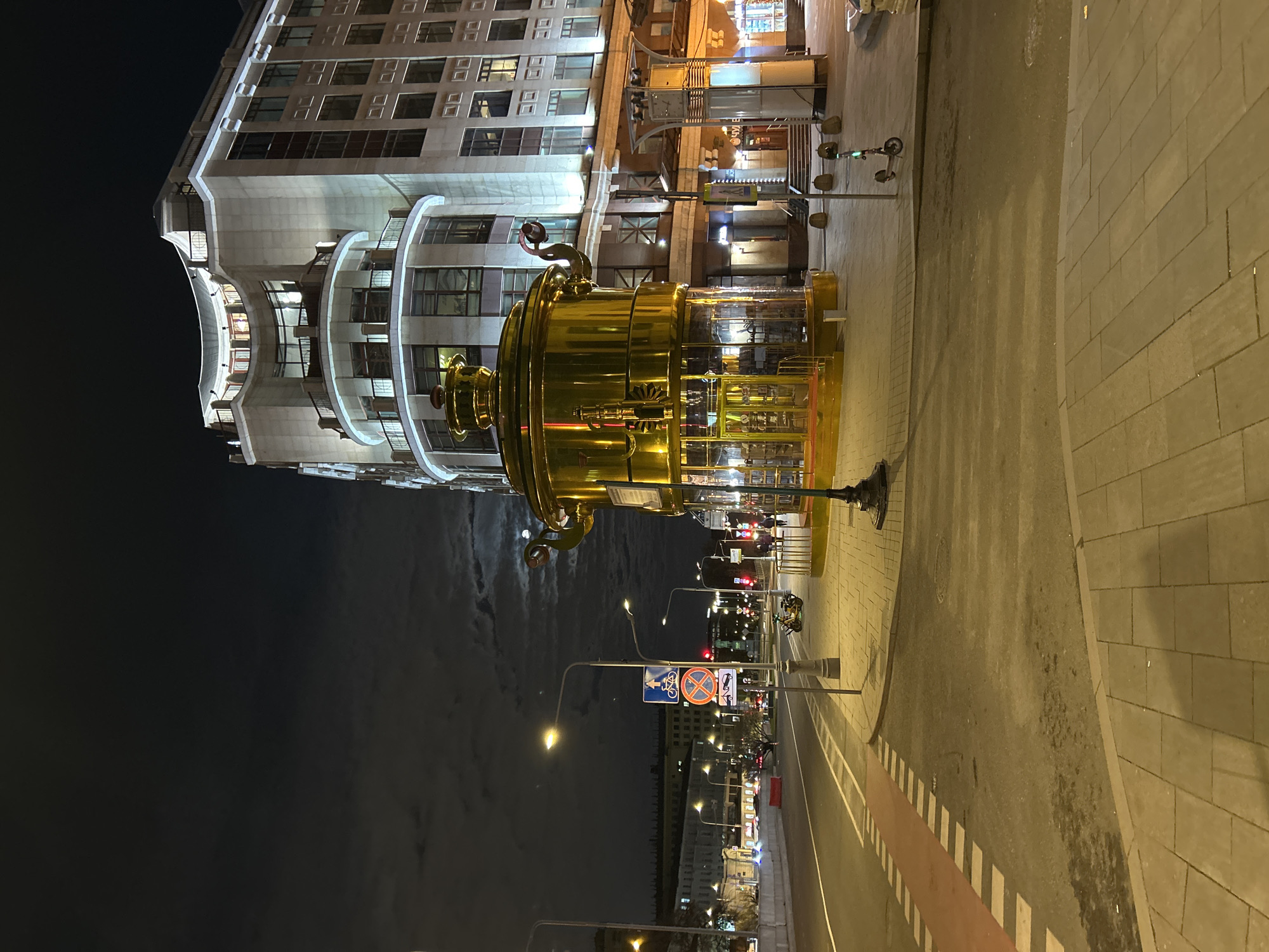 The image shows a street scene at night in an urban area, likely the city of Moscow, Russia.
In the foreground, a large, ornate golden structure resembling a samovar stands prominently on the sidewalk. The building behind the samovar has a light-colored facade with multiple windows and curved architectural features. Streetlights illuminate the street and sidewalk, and traffic signs are visible. Cars drive down the street. The night sky is dark with partial cloud cover, and the moon can be seen in the sky.