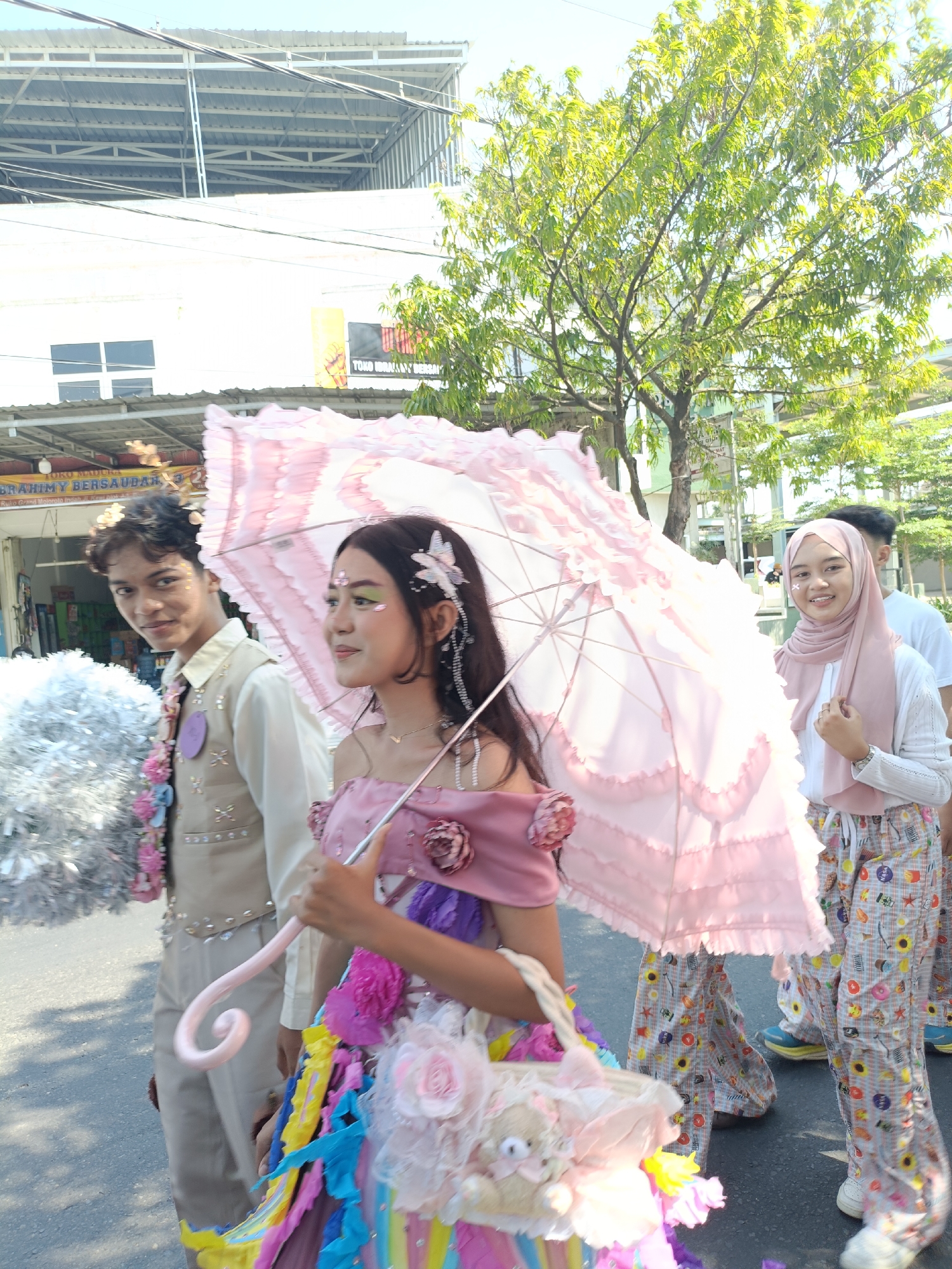 The image shows a street scene in Kediri City, Indonesia, likely during a festival or parade. In the foreground, a young woman in a pink off-the-shoulder dress and an elaborate, ruffled pink umbrella is walking. She has a decorative basket filled with stuffed animals and flowers at her side. Beside her, a young man in a light-colored vest and collared shirt with floral accents looks towards the camera. Further back, another young woman wearing a pink hijab and patterned pants smiles at the camera. Other people and a tree are visible in the background, along with buildings and overhead wires. The scene appears to be outdoors under a bright sky, suggesting daytime.