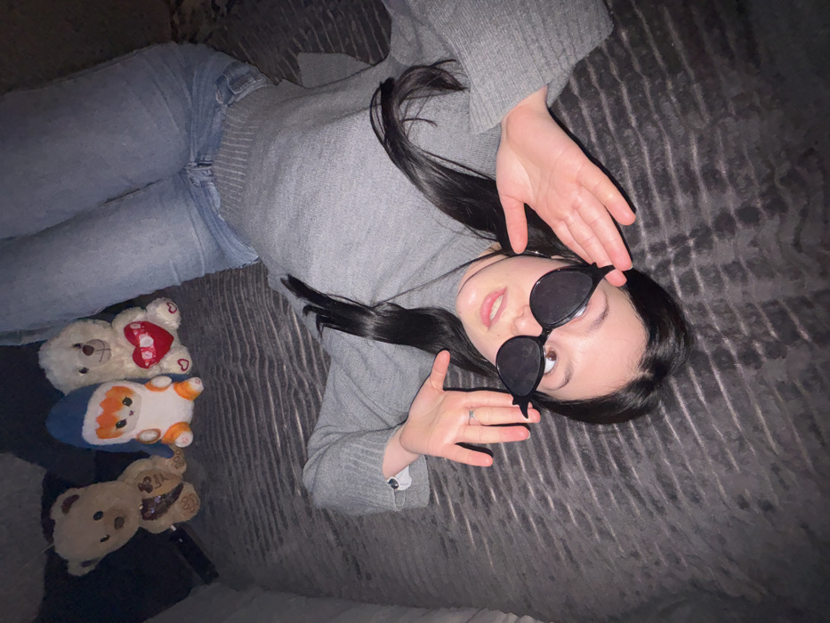 An individual, identified as a young female, is depicted lying supine on a dark grey, horizontally textured surface, likely a bed or sofa, in an indoor setting. She wears a grey long-sleeved sweater and light blue jeans, with her legs extended towards the upper right. Her head is positioned towards the bottom center, tilted slightly right, with her gaze directed towards the viewer. Dark, stylized sunglasses with an upward-swept outer edge rest on her nose, revealing her eyes above the frames. Her hands are open and positioned on either side of her head. A plain metallic ring is visible on the ring finger of her left hand. In the upper left quadrant of the image, three plush toys are arranged: a light brown teddy bear, a white and orange character with a blue hood, and a white teddy bear holding a red heart. The background is indistinct and dimly lit. The specific location within the city of Фоки, Russia, is not discernible from the image.