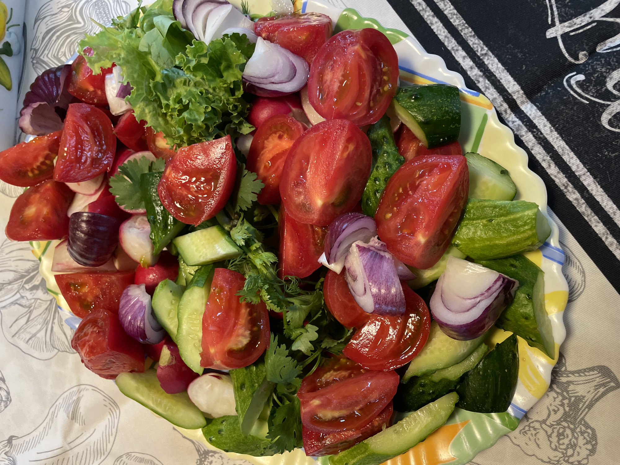 This is a top-down, close-up shot of a colorful salad in a decorative, scalloped-edge bowl. The salad is a mixture of chopped fresh vegetables, including bright red tomatoes, green cucumbers, white radishes, red onion wedges, and leafy green lettuce. Some sprigs of cilantro are also visible, adding a touch of green herb. The vegetables are cut into various shapes and sizes, with tomatoes in wedges and halves, cucumbers in rounds and lengthwise slices, and onions in quartered wedges. The salad appears to be freshly prepared and glistening with moisture, suggesting it is ready to be served. The bowl is resting on a tablecloth with a delicate, light-colored floral or botanical pattern. To the right of the bowl, a black fabric with white stripes and cursive lettering is partially visible, hinting at a placemat or napkin. The lighting is bright, indicating it might be daytime, and there are no people or pets in the frame. The overall impression is one of vibrant, healthy food.