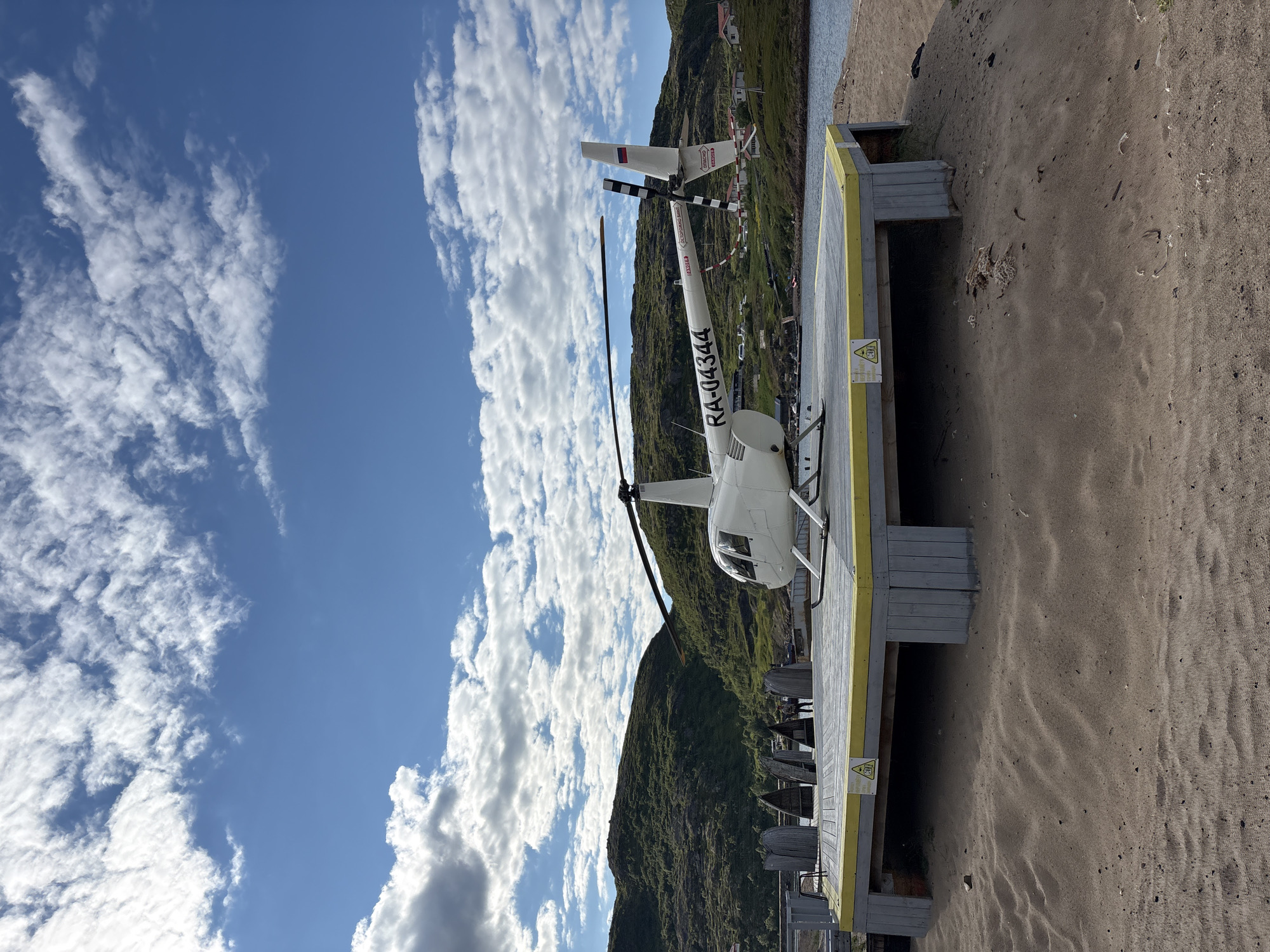A white helicopter with the registration number RA-04344 is parked on a wooden helipad on a sandy embankment in Teriberka, Russia. The helipad is supported by gray wooden posts and has a yellow border. Behind the helipad, a body of water is visible, with small boats docked in the distance. Rolling green hills with sparse vegetation rise behind the water, and scattered houses are nestled on the slopes. The sky is blue with scattered white clouds. The overall scene depicts a remote, coastal location.