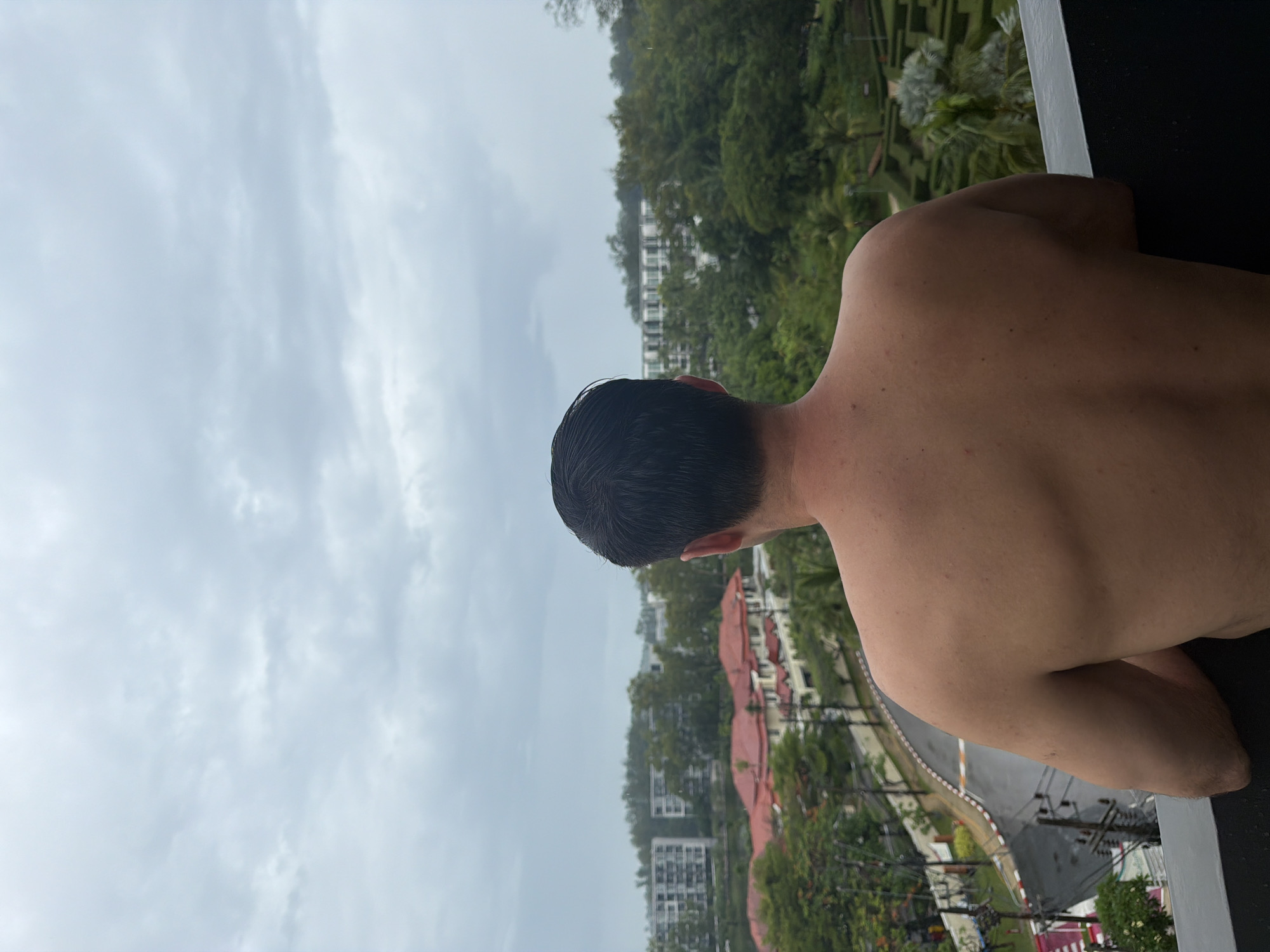 A shirtless, light-skinned man with short, dark hair is viewed from behind and slightly to the right. The man is on a balcony, leaning on a dark surface. The background reveals a landscape of buildings, trees, and a curved road, all under an overcast sky. The image is rotated horizontally. The location is Choeng Thale, Thailand.