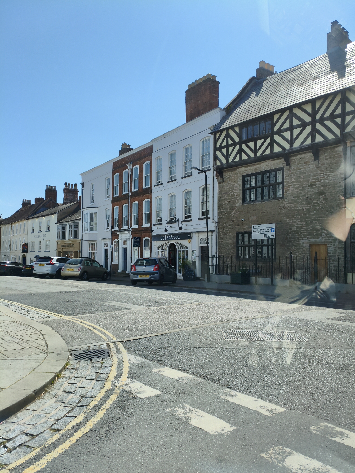 The image shows a street scene in Ludlow, United Kingdom, on a sunny day. The view is from a vehicle, with reflections visible on the windshield. On the right side of the image, a building with a stone lower facade and timber-framed upper stories is prominent. This building has a dark tiled roof and a wooden door. To its left, a row of attached buildings with white and red brick facades extends down the street. These buildings have multiple stories with numerous windows. Several cars are parked along the street. In the foreground, a road with white pedestrian crossing lines and double yellow lines is visible. A curved paved area with cobblestones and a drain cover is on the left. The sky is clear and blue.