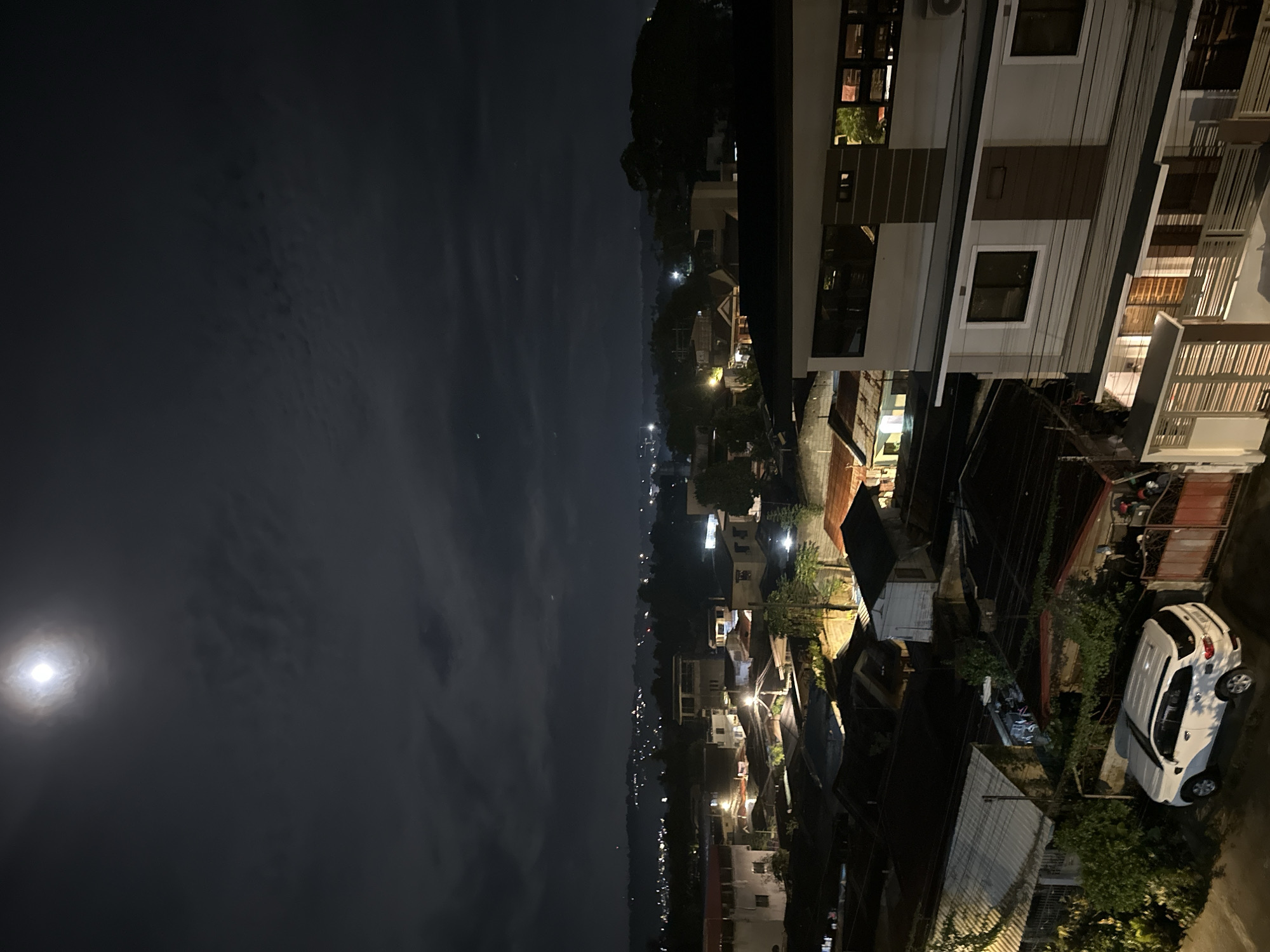 The image captures a night scene in a residential area of Davao City, Philippines, under a cloudy sky illuminated by a bright full moon. The foreground features a two-story building with a modern facade, characterized by neutral colors and horizontal accent lines. Below this building, a white SUV is parked on a paved surface adjacent to a metal gate and lush greenery.
The midground reveals a dense collection of houses with various roof types, predominantly dark-colored, and a mix of architectural styles, indicating a well-established neighborhood. Streetlights and lights from within the houses create a warm glow amidst the darkness.
The background extends to a sprawling cityscape with numerous lights dotting the landscape, suggesting a broader urban area with rolling hills or mountains in the distance. The overall impression is one of a tranquil, lived-in community at night, bathed in the ethereal light of the moon.