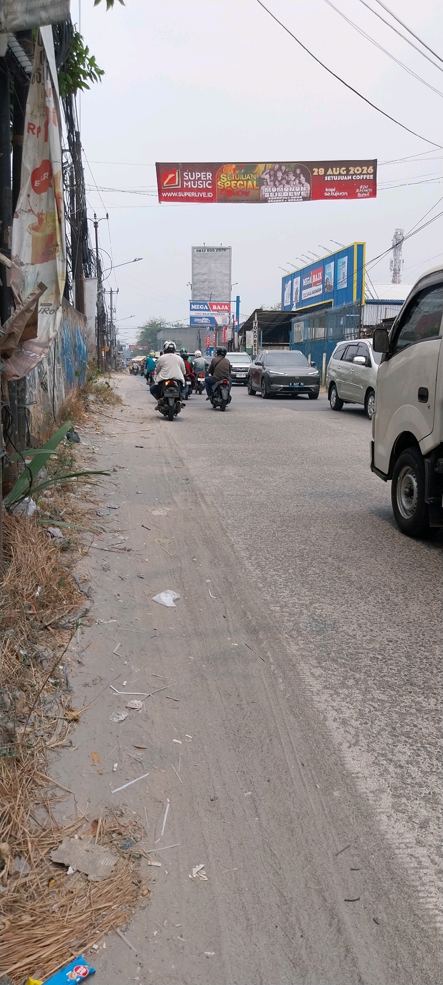 The image depicts a street scene in Bojong Nangka, Indonesia, likely on a cloudy day, given the overcast sky. The foreground is dominated by a dirt and gravel road, littered with dried vegetation, twigs, and various pieces of trash, including plastic bags and cardboard. The left side of the road features a grassy embankment with more debris. Power lines and poles are prominently visible overhead, with a few leafy branches extending from the left.
In the middle ground, several motorbikes with riders are navigating the road. The riders are wearing helmets and casual clothing. To the right, several cars are present, including a silver SUV and a darker colored vehicle, along with a white truck partially visible on the far right. A few buildings or structures are visible in the background, including what appears to be a blue building with yellow trim.
Two large banners are suspended over the street. One banner, primarily red and brown, advertises "SUPER MUSIC SETUJUAN" and "MOMONON SEJEDEWE" with a date of "28 AUG 2026" and "SETUJUAN COFFEE." Another billboard, to the left of this banner, displays "MEGA BAJA" and "HEMAT BAJA INDONESIA." A smaller, white billboard with a phone number is also visible further down the street. The overall impression is of a busy, somewhat unkempt urban or suburban street.