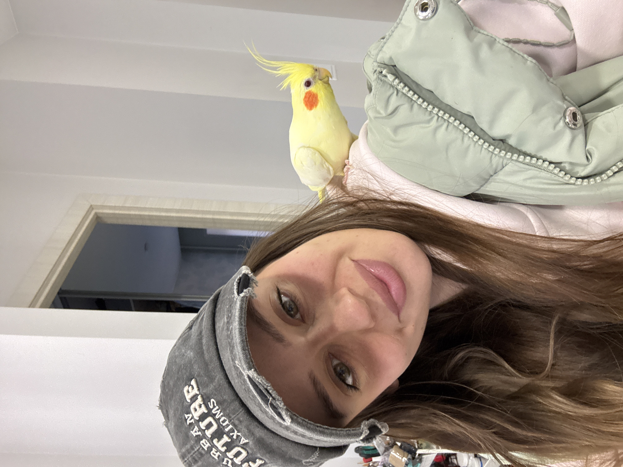 A woman with long brown hair and a gray cap labeled "URBAN FUTURE" is taking a selfie. A yellow cockatiel with orange cheek patches is perched on her shoulder. She wears a light green puffer jacket over a pink shirt. The location appears to be indoors, based on the door frame and plain white walls, but the exact location in Krasnoyarsk, Russia, is not discernible from the image alone.