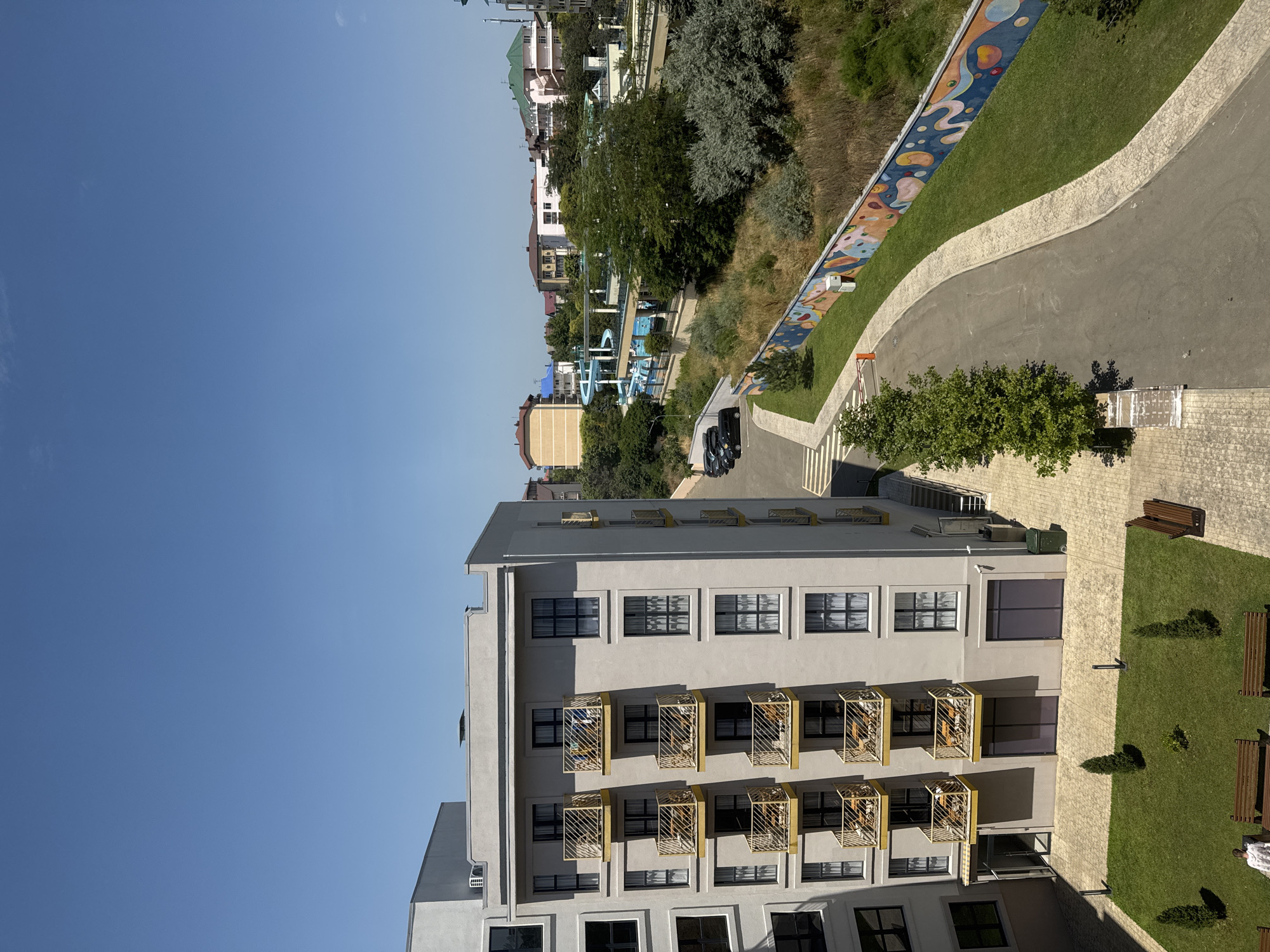 The image depicts a view of a modern apartment building with balconies, an adjacent courtyard with benches and a grassy area, and a street curving along a colorful mural-decorated wall. Beyond the building, a glimpse of a water park with slides and pools is visible, along with other buildings and trees characteristic of a coastal resort town. The sky is clear blue. The scene appears to be in Vityazevo, Russia, a resort town known for its beaches and entertainment facilities.