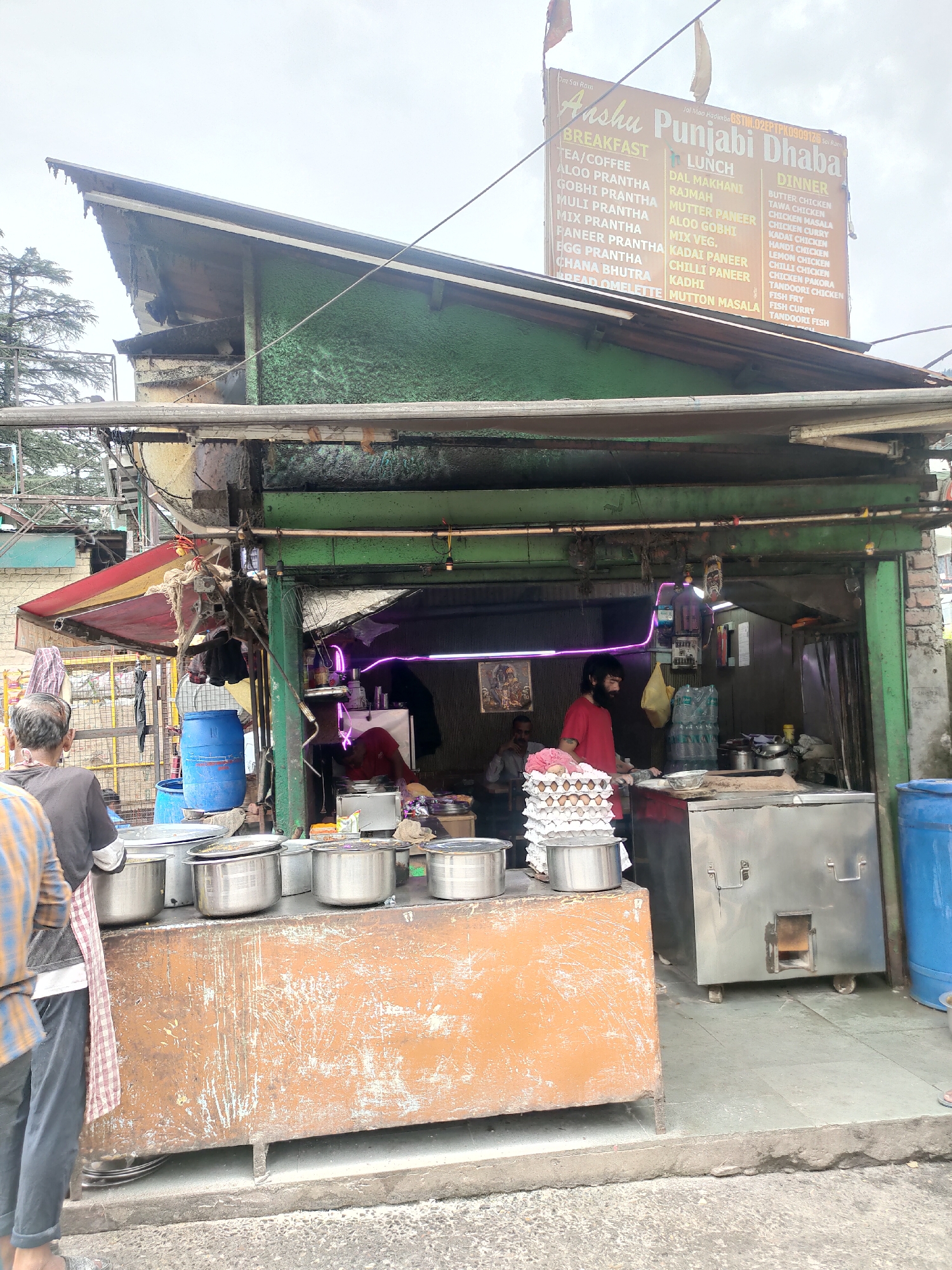 The image depicts a street-side food stall, identified as "Anshu Punjabi Dhaba," situated in Manali, India. The stall features a prominent menu board listing breakfast, lunch, and dinner items, including various parathas, dals, and chicken dishes. Two individuals are visible in the foreground: one man in a black shirt and another in a blue and yellow plaid shirt, both appearing to be customers or staff. Inside the stall, a person in a red shirt is actively preparing food. Various cooking utensils, pots, and an egg carton are arranged on a counter. Behind the counter, a large stainless steel tandoor oven and a blue barrel are present. The stall has a makeshift roof and appears to be open-air, with a backdrop of trees and buildings suggesting an urban or semi-urban environment. The overall scene conveys a bustling, everyday scene of food service in a popular tourist destination.