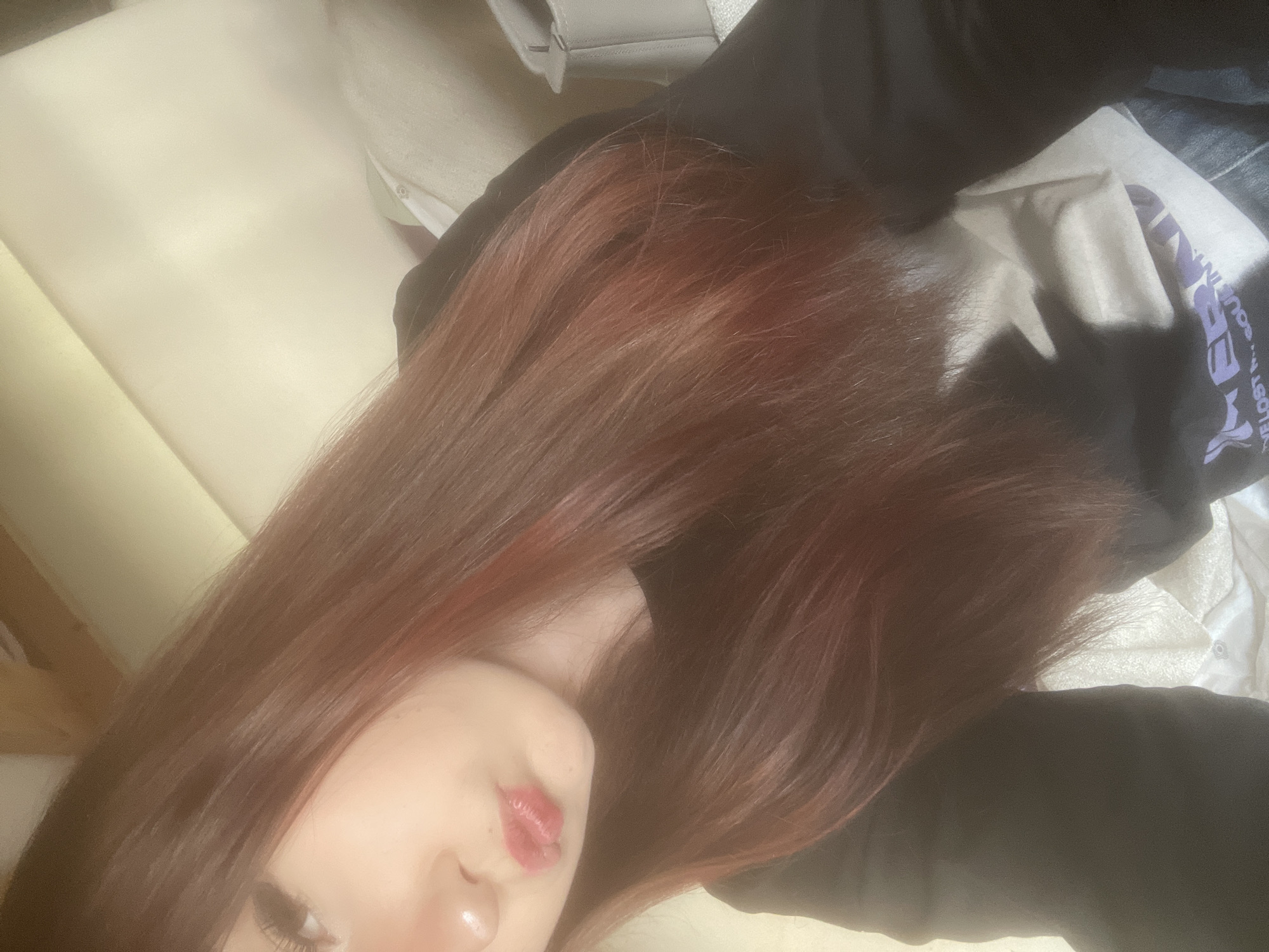 This close-up image captures a person, likely a young woman, reclining in an indoor setting. The photograph is oriented 90 degrees counter-clockwise. The prominent feature is her long, wavy reddish-brown hair, which flows across most of the frame.
Only the lower left side of her face is visible, including her left cheek, part of her nose, and her lips, which are painted with a glossy reddish-pink lipstick and form a subtle pout. She has light skin and visible dark eyelashes. Her expression appears relaxed.
She is wearing a dark garment, possibly black, which covers her body on the right side of the image. On this dark fabric, fragments of purple text are visible, reading "WE LOST SOUL...", suggesting it might be part of a graphic on her clothing. A crumpled white tissue or paper is also seen on the dark fabric. In the background, a light-colored, possibly cream or off-white, surface suggests a pillow, bed, or couch. A light grey object with a metallic detail is briefly visible in the upper background, potentially a bag or part of the furniture. The lighting is soft and ambient, indicative of an indoor setting during the day. No specific visual cues within the image confirm the provided location context of Saint Petersburg, Russia.