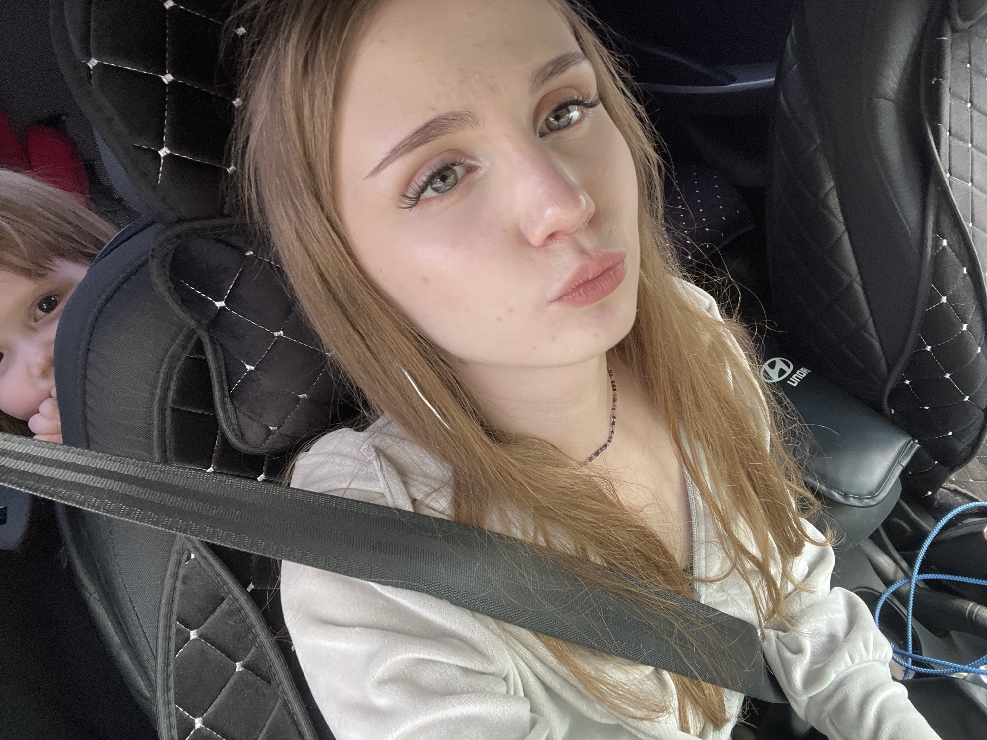The image shows a young woman and a child in a car. The woman is in the foreground, looking at the camera with her mouth puckered. She has long, blonde hair and is wearing a light-colored shirt and a necklace. A seatbelt is visible across her chest. To her left, partially visible, is a child with blonde hair, looking towards the camera. The background consists of car seats, one of which is black with diamond-patterned stitching. There is also a glimpse of a blue cord on the right side of the frame. The scene appears to be inside a vehicle, possibly a car, and is not specifically located in any part of Новая Отрадовка, Russia, as there are no identifying landmarks or indicators of that location.