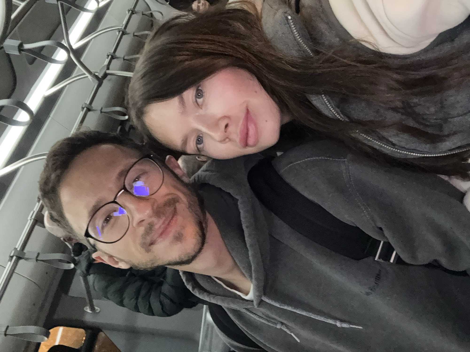 The image displays two individuals, an adult male and an adult female, captured in a close-up selfie within the interior of a public transport vehicle, consistent with a metro train on the Athens Metro system, which serves the Paiania Municipal Unit, Greece. The male, positioned on the left, has short brown hair, a short beard, and wears black-framed glasses, a grey hooded sweatshirt, and a black backpack strap. He presents a slight smile while looking directly at the camera. The female, on the right, possesses long dark brown hair, light-colored eyes, and wears a grey zippered jacket over a light-colored garment. Her gaze is directed towards the camera with a neutral expression. The overhead environment features numerous grey strap handles attached to metal bars and a horizontal fluorescent light fixture. A blurred individual in a dark puffer jacket is partially visible behind the male, and another obscured figure is in the distant background on the right. An orange light source is visible through a window in the upper left, indicating an external environment, likely at dusk or night.