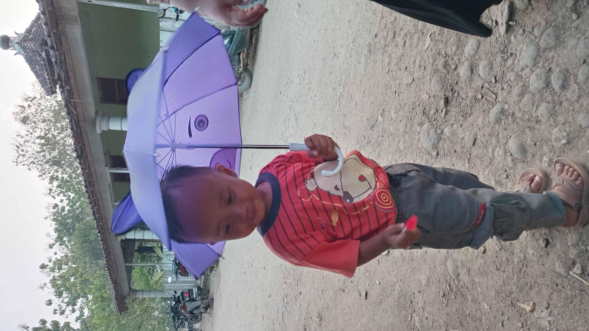 A young boy stands outdoors in Kedungwaru, Indonesia, holding a purple umbrella with a cartoon face. He is wearing a red and black striped t-shirt with a bear graphic and gray cargo pants. The boy is also wearing brown sandals. The background shows a building with a tiled roof, trees, and motorcycles parked on a dirt road. The sky is overcast.