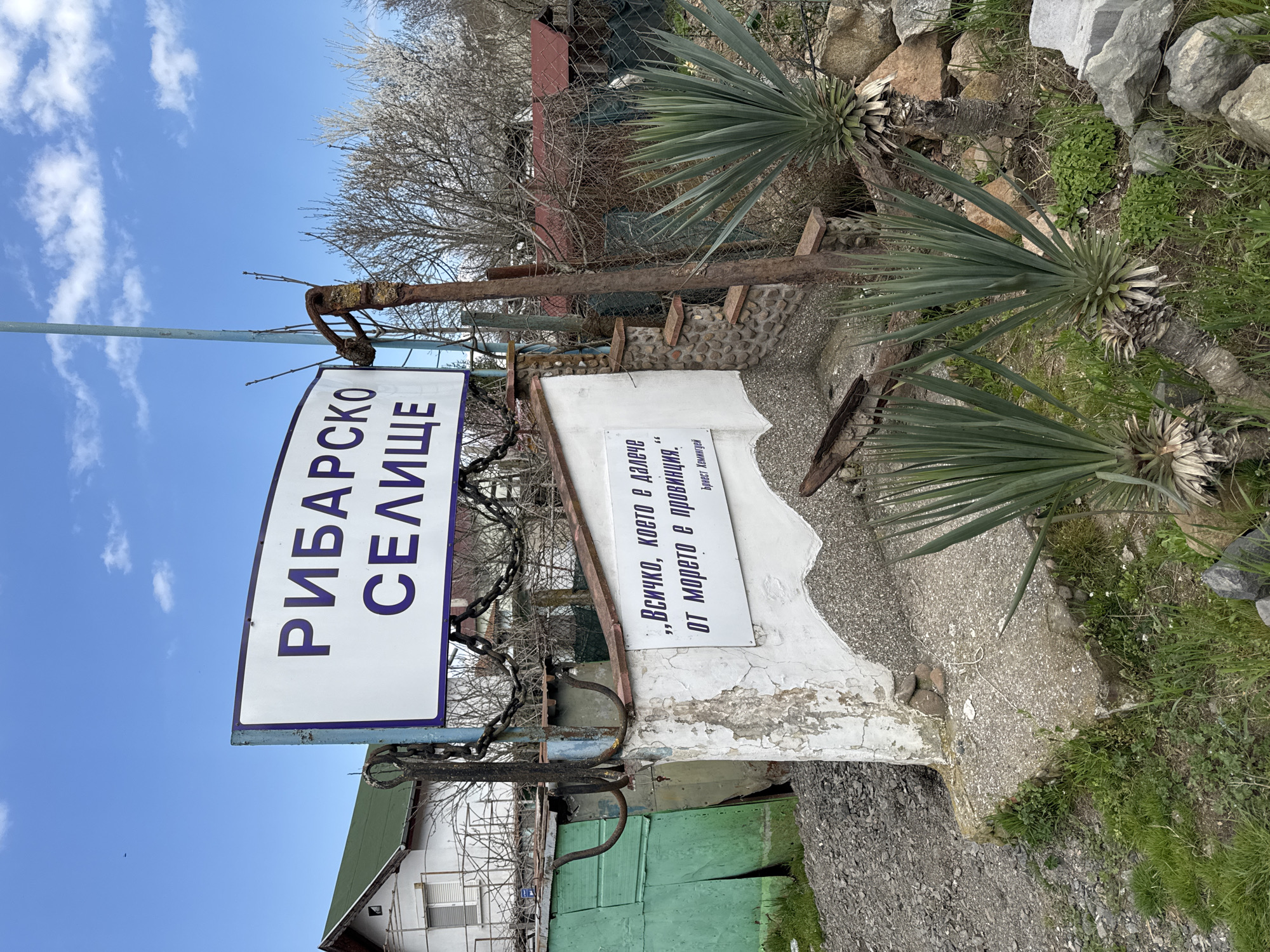 A slightly tilted shot depicts an outdoor area with a white sign displaying the text "РИБАРСКО СЕЛИЩЕ" in bold, blue Cyrillic letters. Below, a smaller sign contains another Cyrillic quote attributed to "Ърнест Хемингуей" (Ernest Hemingway).
The setting is somewhat rustic, with a weathered white wall and gray concrete pathway visible. Plants with sharp, green fronds grow in the area next to a rocky patch. Faded green paint is seen on a structure behind the sign. A metal fence and bare trees are in the background, under a clear blue sky with a few white clouds. The scene appears to be in Burgas, Bulgaria.
