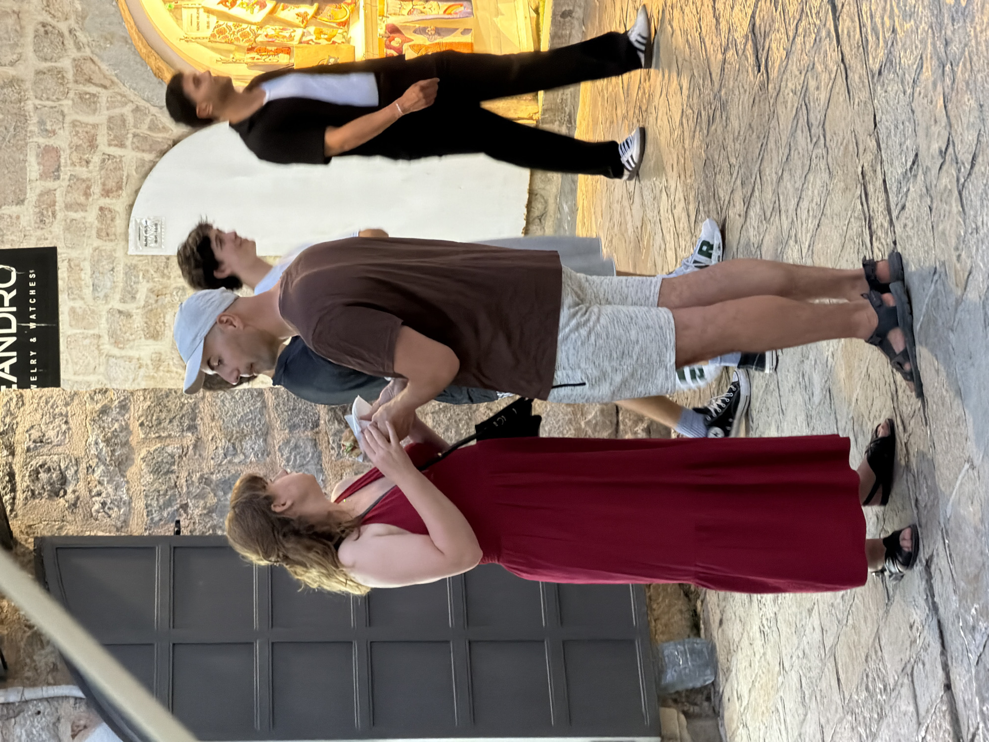 In Budva, Montenegro, on an outdoor cobblestone street, a group of people are interacting and walking. A woman in a red maxi dress and black sandals holds a white paper object and appears to be speaking with a man wearing a grey baseball cap, a brown t-shirt, and grey shorts with black sandals. Behind them, a young man in a white t-shirt and a man in a black short-sleeved shirt and black pants are also visible. The background features a stone wall and an arched opening leading to a shop with merchandise displayed. The lighting suggests it is late afternoon or early evening.