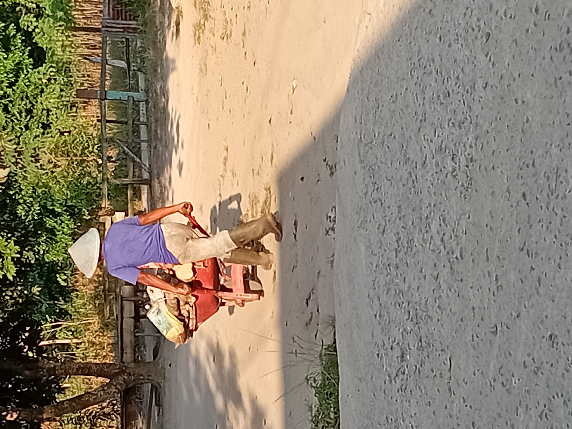 In Kasang Pudak, Indonesia, a person wearing a conical straw hat and a purple t-shirt is pushing a red wheelbarrow. The wheelbarrow is loaded with what appears to be building materials, including bags of cement or similar powder, and possibly some wooden items. The individual is also wearing light-colored pants and dark boots. The scene is outdoors on a dirt or gravel path, with vegetation and a wooden fence visible in the background. The lighting suggests it is daytime, possibly late afternoon, with a shadow cast on the ground.
