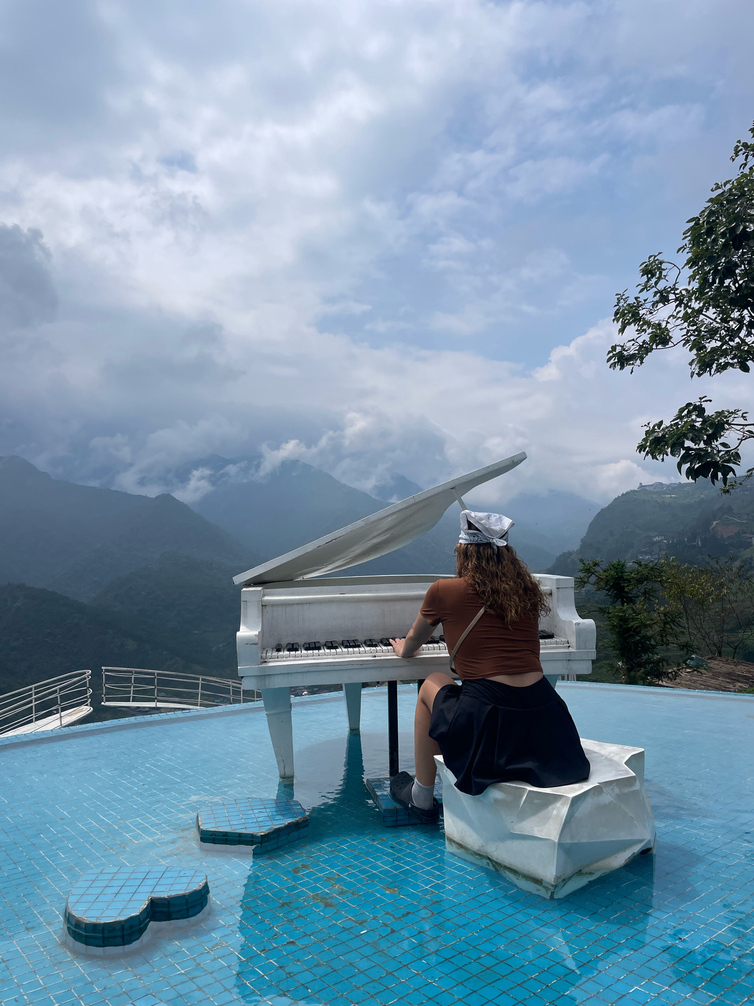 A person with curly brown hair, wearing a brown t-shirt, black skirt, white bandana on their head, and black Crocs, is playing a white grand piano. The piano is positioned on a blue tiled surface that appears to be a pool or a reflective surface, surrounded by water. In front of the piano and to the left, there are two abstract, blue-tiled structures rising from the water, resembling musical notes. The person is seated on a geometric, white stool. Behind the piano and the person, a vast mountainous landscape is visible, shrouded in mist and clouds. The mountains are covered in lush green vegetation. In the upper right corner of the image, a portion of a tree with green leaves is visible. The overall scene suggests an outdoor setting, possibly a viewpoint or an attraction in Sa Pa Ward, Vietnam, offering a unique juxtaposition of music and natural scenery.