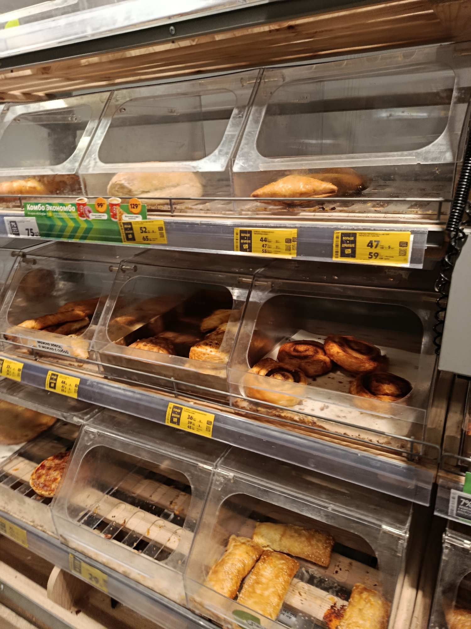 The image shows a close-up, eye-level view of baked goods displayed in a supermarket bakery section in Vareya, Russia. The baked goods are housed in clear plastic bins, organized on multiple shelves. The lighting is bright and appears to be from overhead sources, typical of a retail environment. On the top shelf, a long white bread loaf and a golden-brown pastry are visible. The middle shelves contain several types of pastries, including what appear to be filled buns or empanadas, and cinnamon rolls. The bottom shelf displays smaller, rectangular pastries, some with sesame seeds. Price tags and promotional signs are affixed to the shelves, some in Russian. The overall environment is clean and orderly, suggesting a well-maintained retail space. No people are visible in the image. The context is that of a commercial establishment selling food items.