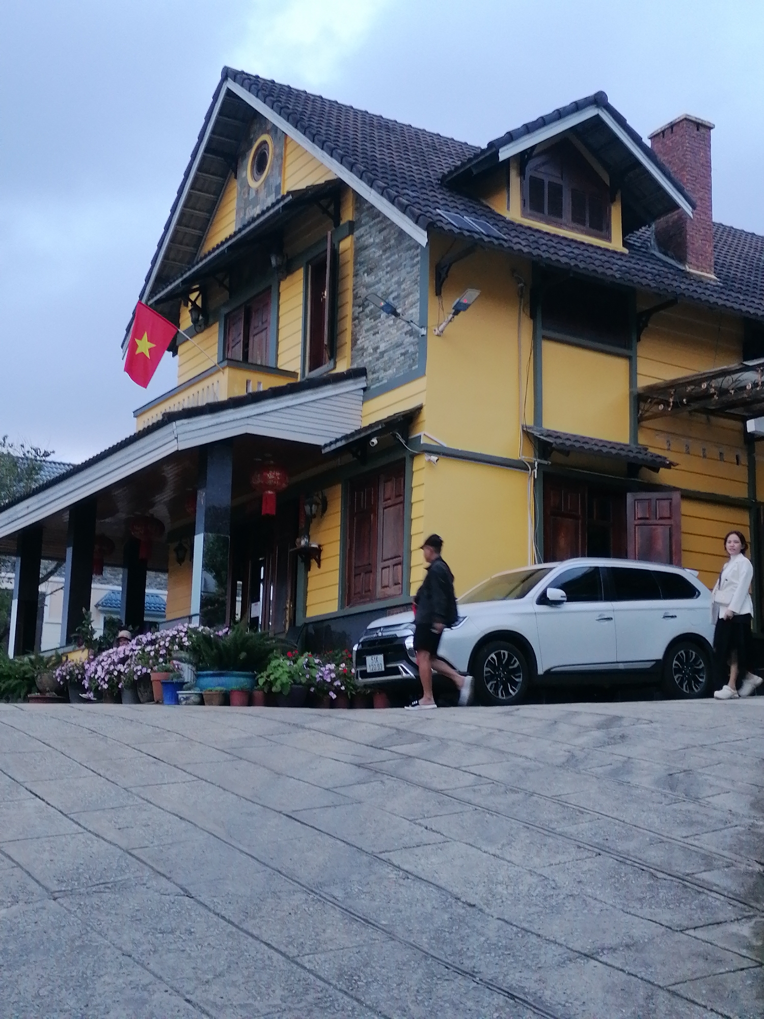 The image depicts a scene in Xuân Trường - Đà Lạt, Vietnam. In the foreground, a gray, paved pathway slopes upwards towards a yellow building with a dark tiled roof. The building features traditional wooden shutters on its windows and a prominent red brick chimney. A Vietnamese flag with a yellow star is visible flying from a pole on the left side of the building.
In front of the building, a white SUV is parked. Two individuals are present near the vehicle: a person wearing a dark jacket and shorts is walking away from the car, and another person in a white jacket and black skirt is standing on the right side of the car, facing away from the viewer. A line of potted plants, including pink and purple flowers, adorns the area in front of the building. The sky is overcast, suggesting a potentially cool or damp day.
