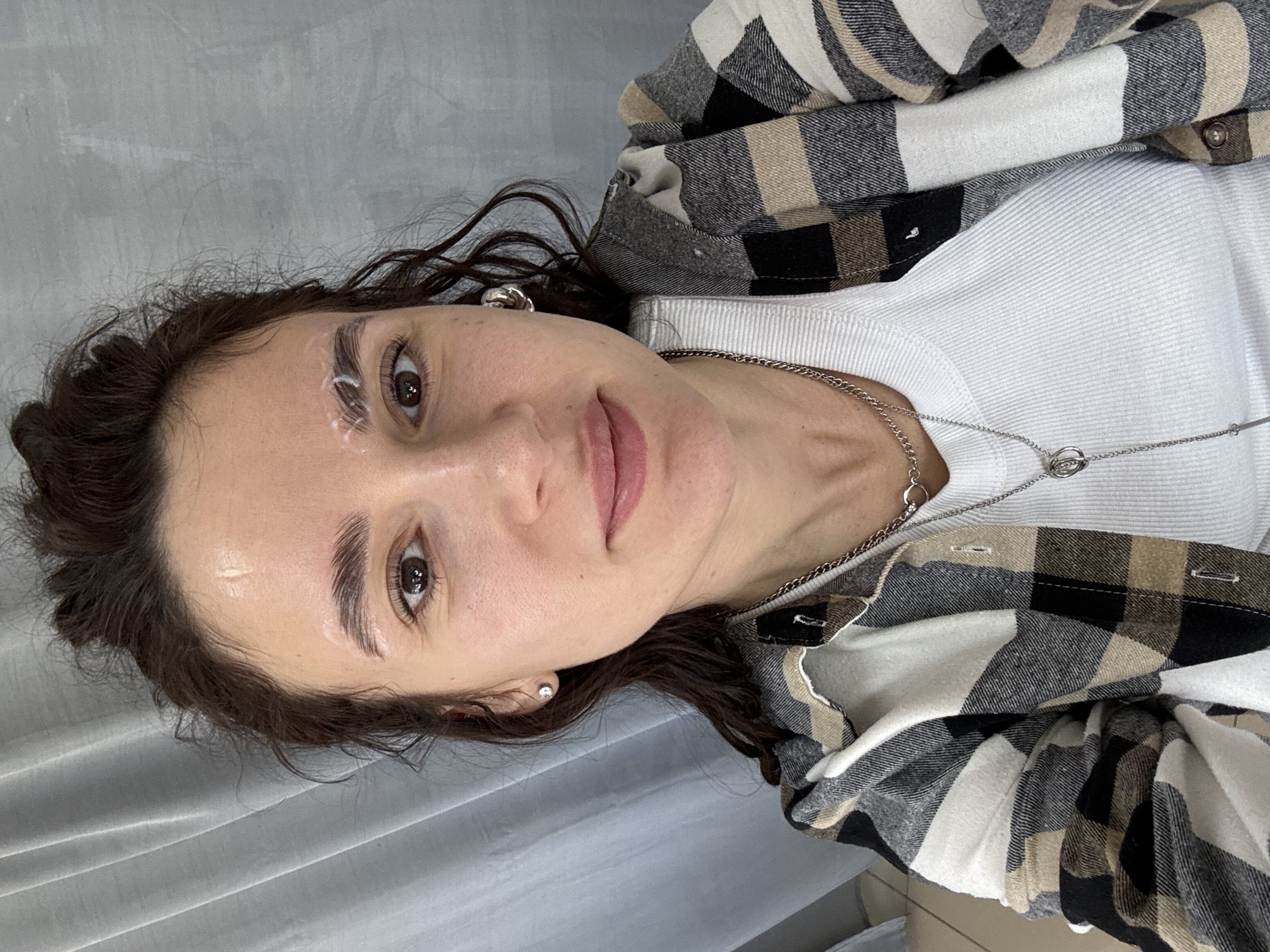 A young woman is depicted in a close-up portrait, lying down and looking directly at the camera with a neutral to slightly smiling expression. She has dark, wavy hair, brown eyes, and fair skin.
A notable detail is the white cream or paste applied to both of her eyebrows, indicating she is undergoing a cosmetic beauty treatment, such as brow lamination, tinting preparation, or mapping for a permanent makeup procedure.
She is dressed in casual layered clothing, consisting of a white ribbed long-sleeved top worn underneath an open plaid flannel shirt. The flannel shirt features a pattern of black, white, and tan or beige squares. A silver chain necklace with a small, intertwined circular pendant is visible around her neck, and she wears small, clear stud earrings.
The background is a plain, light grey surface, possibly a textured wall or a soft fabric curtain, suggesting an indoor setting like a beauty salon or spa. The overall lighting is soft and even. No specific time of day can be determined, and there is no visible text in the image. The setting is in Chaikovsky, Russia.