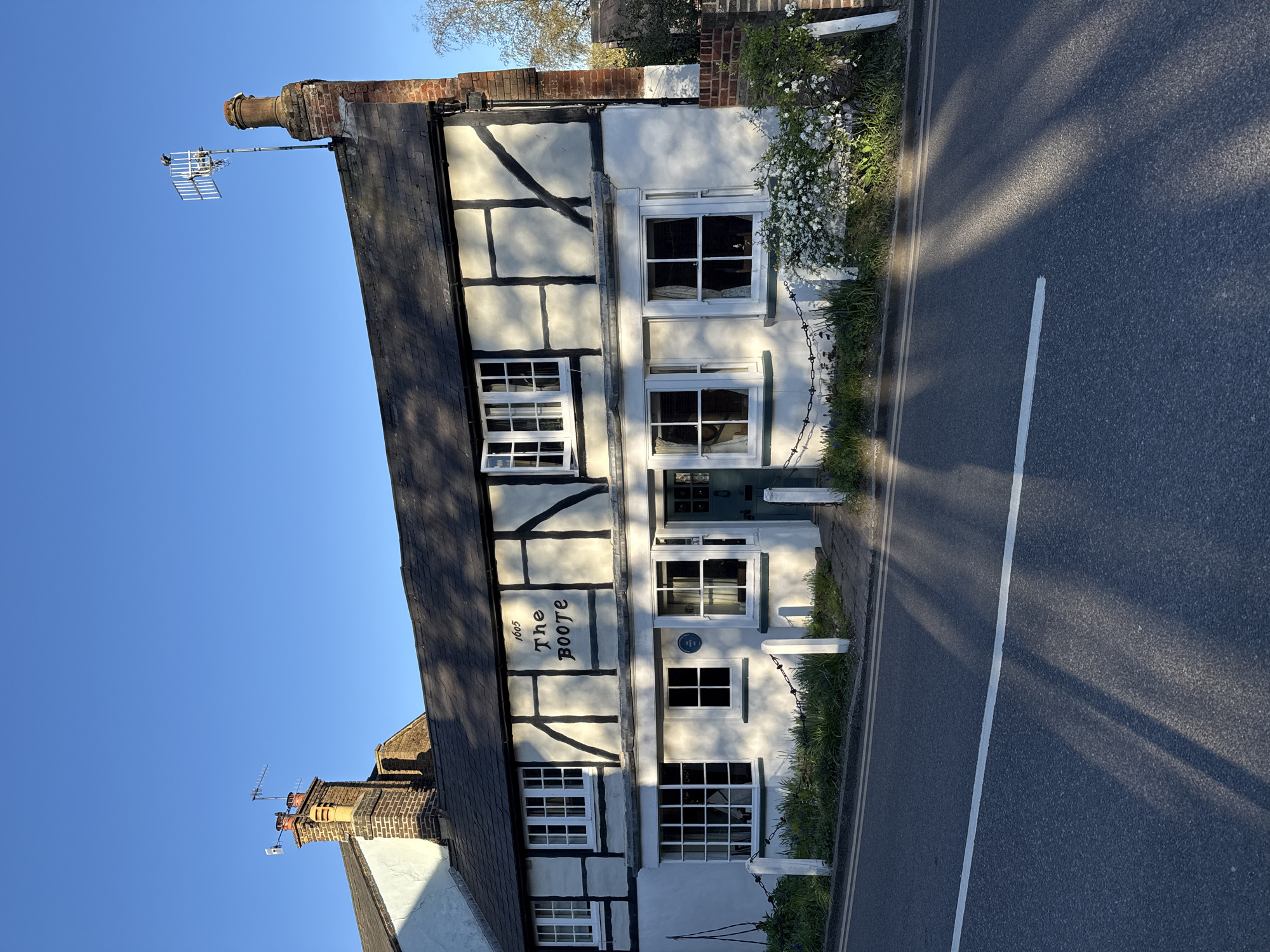 The image shows a historic building with a half-timbered facade, located on a street in Dacorum, United Kingdom. The building has the name "The Boote" and the year "1605" painted on its facade. It has multiple windows with white frames and a blue door. The building is situated next to a paved road with a white line marking. A chain-link fence separates the building from the road. Vegetation grows along the base of the building and the edge of the road. A television antenna is visible on the roof. The sky is clear and blue.